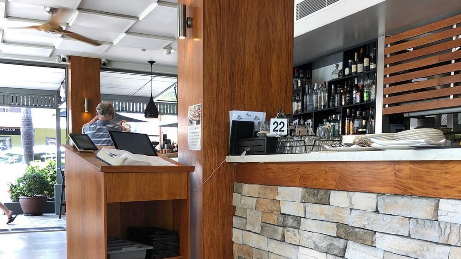 On the left, a wooden host counter with a register, papers, and a storage shelf below. A man in a striped shirt stands near glass doors, with outdoor seating visible beyond. In the middle, a wooden column extends from floor to ceiling. On the right, a stone-fronted counter topped with white marble holds plates, cutlery, and a numbered sign. Behind it, shelves with rows of bottles and glasses are framed by wooden slats. The floor is tiled, and the white ceiling has recessed panels. On the left, a wooden host counter with a register, papers, and a storage shelf below. A man in a striped shirt stands near glass doors, with outdoor seating visible beyond. In the middle, a wooden column extends from floor to ceiling. On the right, a stone-fronted counter topped with white marble holds plates, cutlery, and a numbered sign. Behind it, shelves with rows of bottles and glasses are framed by wooden slats. The floor is tiled, and the white ceiling has recessed panels.