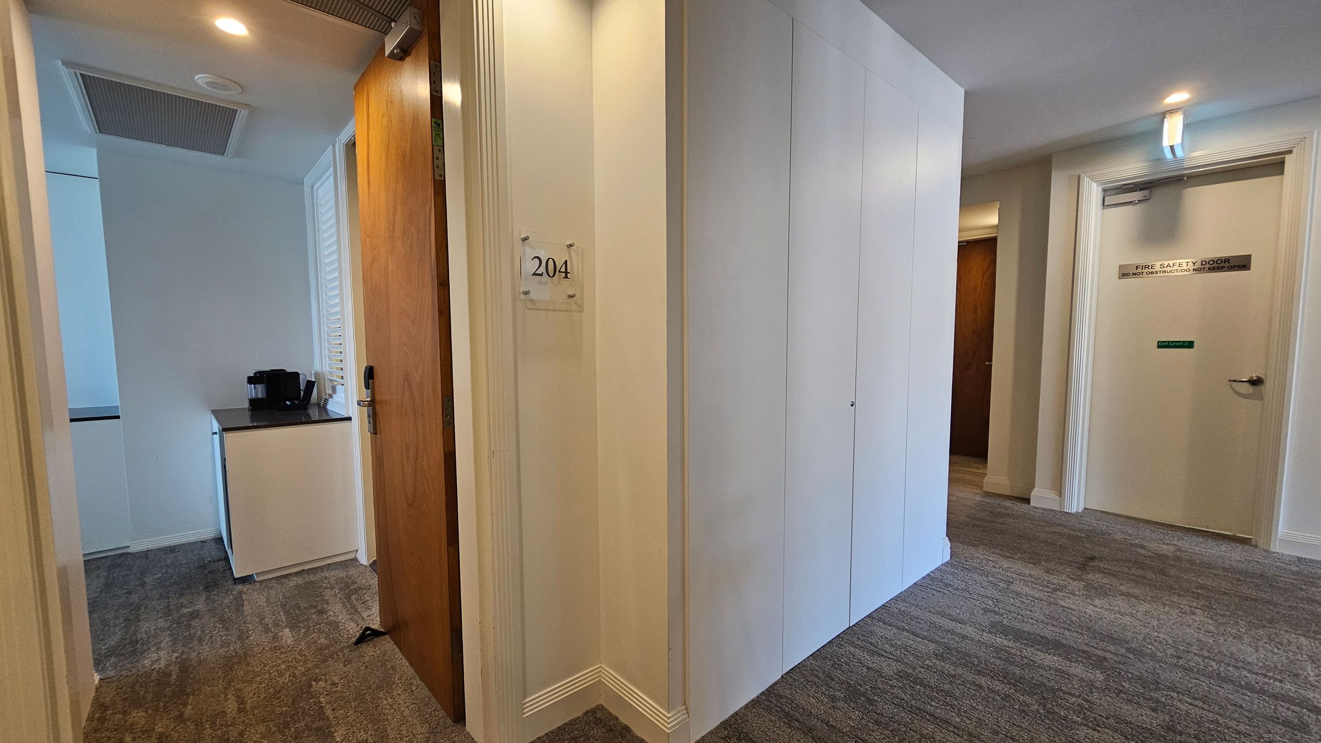 A hallway with a grey and green patterned carpet. On the left, a wooden door is slightly open, revealing a small room with a black coffee machine on a white cabinet. The door has a sign with the number "204". On the right, a white door marked "Fire Safety Door" is closed. The walls are light-coloured, and the ceiling is white with recessed lighting. A hallway with a grey and green patterned carpet. On the left, a wooden door is slightly open, revealing a small room with a black coffee machine on a white cabinet. The door has a sign with the number "204". On the right, a white door marked "Fire Safety Door" is closed. The walls are light-coloured, and the ceiling is white with recessed lighting.