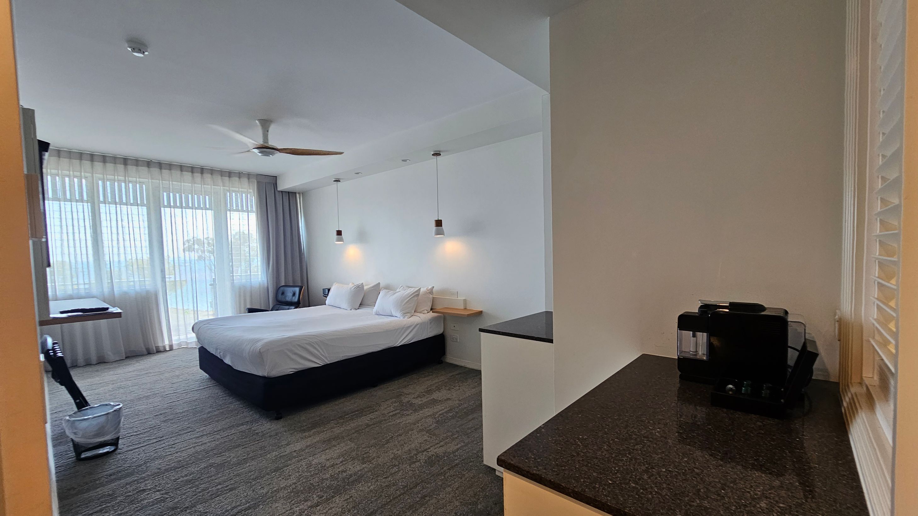 A hotel room with a large bed in the centre, adorned with white bedding and pillows. Two pendant lights hang above the bed. On the left, a large window with sheer curtains allows natural light to enter. A small table and chair are near the window. On the right, a black coffee machine sits on a dark countertop. The floor is carpeted in a grey and green pattern, and a ceiling fan is above the bed. A hotel room with a large bed in the centre, adorned with white bedding and pillows. Two pendant lights hang above the bed. On the left, a large window with sheer curtains allows natural light to enter. A small table and chair are near the window. On the right, a black coffee machine sits on a dark countertop. The floor is carpeted in a grey and green pattern, and a ceiling fan is above the bed.