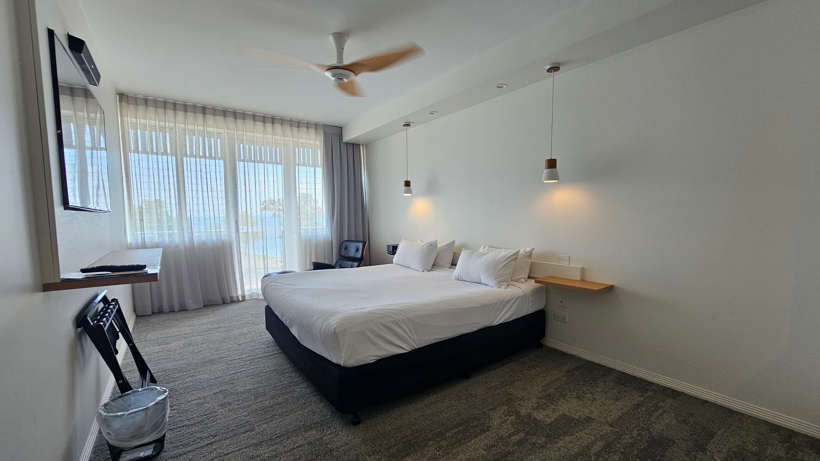 A modern hotel room with a large bed in the centre, adorned with white linens and pillows. Above the bed, two pendant lights hang from the ceiling. On the right, a small wooden shelf is attached to the wall. The left side features a wall-mounted TV above a folding chair and a waste bin. Large windows with sheer curtains allow natural light to fill the room. The floor is carpeted in a mix of grey and green tones, and a ceiling fan is visible above. A modern hotel room with a large bed in the centre, adorned with white linens and pillows. Above the bed, two pendant lights hang from the ceiling. On the right, a small wooden shelf is attached to the wall. The left side features a wall-mounted TV above a folding chair and a waste bin. Large windows with sheer curtains allow natural light to fill the room. The floor is carpeted in a mix of grey and green tones, and a ceiling fan is visible above.