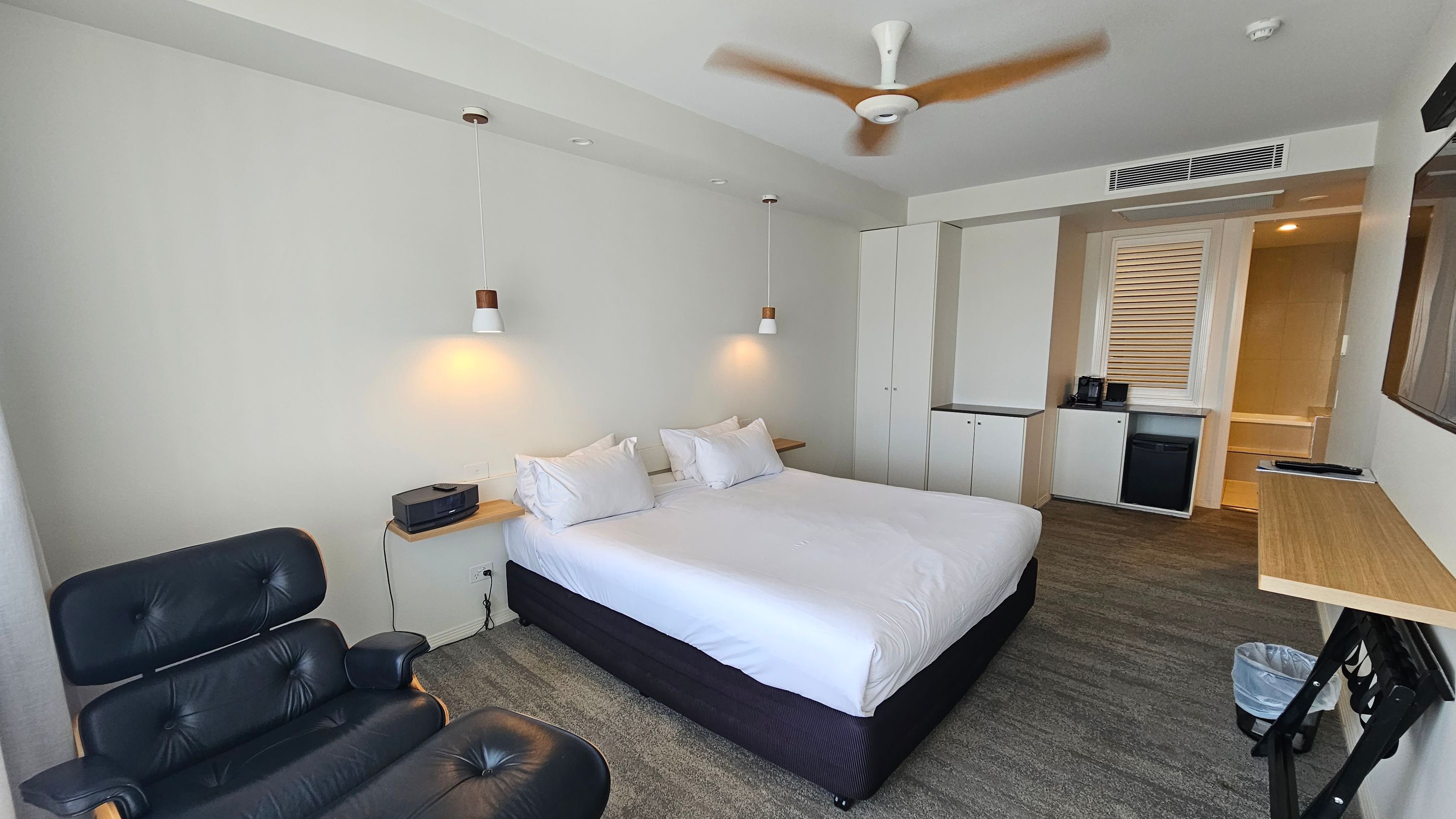 A modern hotel room with a large bed in the centre, flanked by two hanging pendant lights. On the left, there's a black leather chair. To the right, a small kitchenette area with a mini fridge, coffee maker, and a cupboard. A wooden desk is mounted on the wall, with a waste bin underneath. The ceiling features a spinning fan, and the floor is carpeted in a mix of grey and green tones. A modern hotel room with a large bed in the centre, flanked by two hanging pendant lights. On the left, there's a black leather chair. To the right, a small kitchenette area with a mini fridge, coffee maker, and a cupboard. A wooden desk is mounted on the wall, with a waste bin underneath. The ceiling features a spinning fan, and the floor is carpeted in a mix of grey and green tones.
