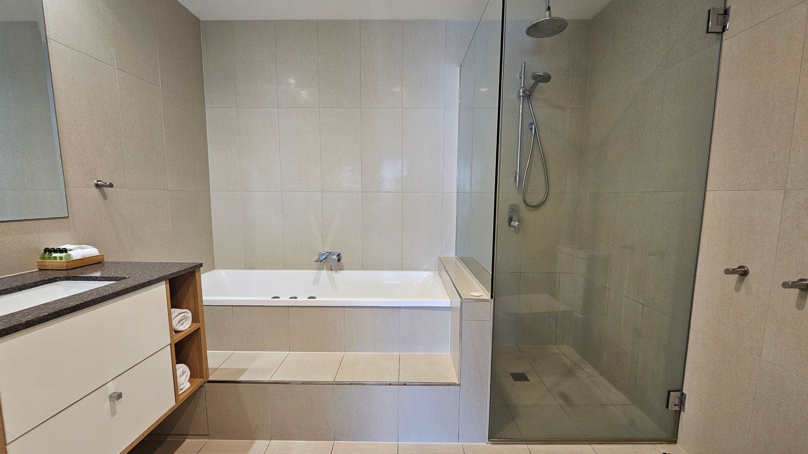 A modern bathroom with a beige tiled floor and walls. On the left, a vanity unit with a dark countertop holds a sink, toiletries, and towels on open shelves. In the centre, a white bathtub is set into a tiled platform. On the right, a glass-enclosed shower area features a rainfall showerhead and a handheld shower. The ceiling is plain white. A modern bathroom with a beige tiled floor and walls. On the left, a vanity unit with a dark countertop holds a sink, toiletries, and towels on open shelves. In the centre, a white bathtub is set into a tiled platform. On the right, a glass-enclosed shower area features a rainfall showerhead and a handheld shower. The ceiling is plain white.