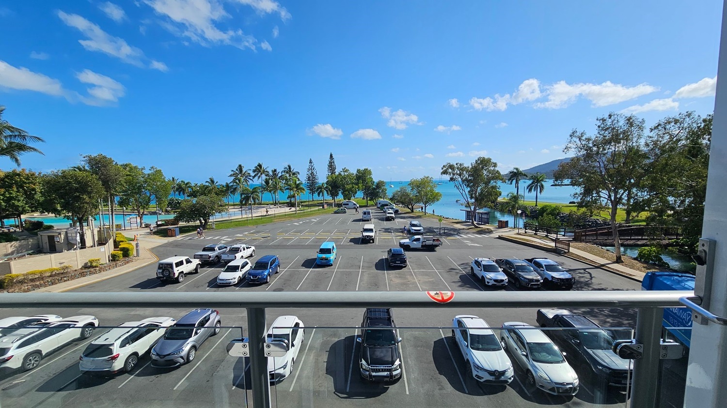A view from a balcony overlooking a car park with various vehicles, including white and blue cars. Beyond the car park, there are palm trees and a pathway leading to a beach with turquoise water. On the right, a small bridge crosses over a stream surrounded by trees. The scene is bright and sunny, with a clear blue sky. A view from a balcony overlooking a car park with various vehicles, including white and blue cars. Beyond the car park, there are palm trees and a pathway leading to a beach with turquoise water. On the right, a small bridge crosses over a stream surrounded by trees. The scene is bright and sunny, with a clear blue sky.