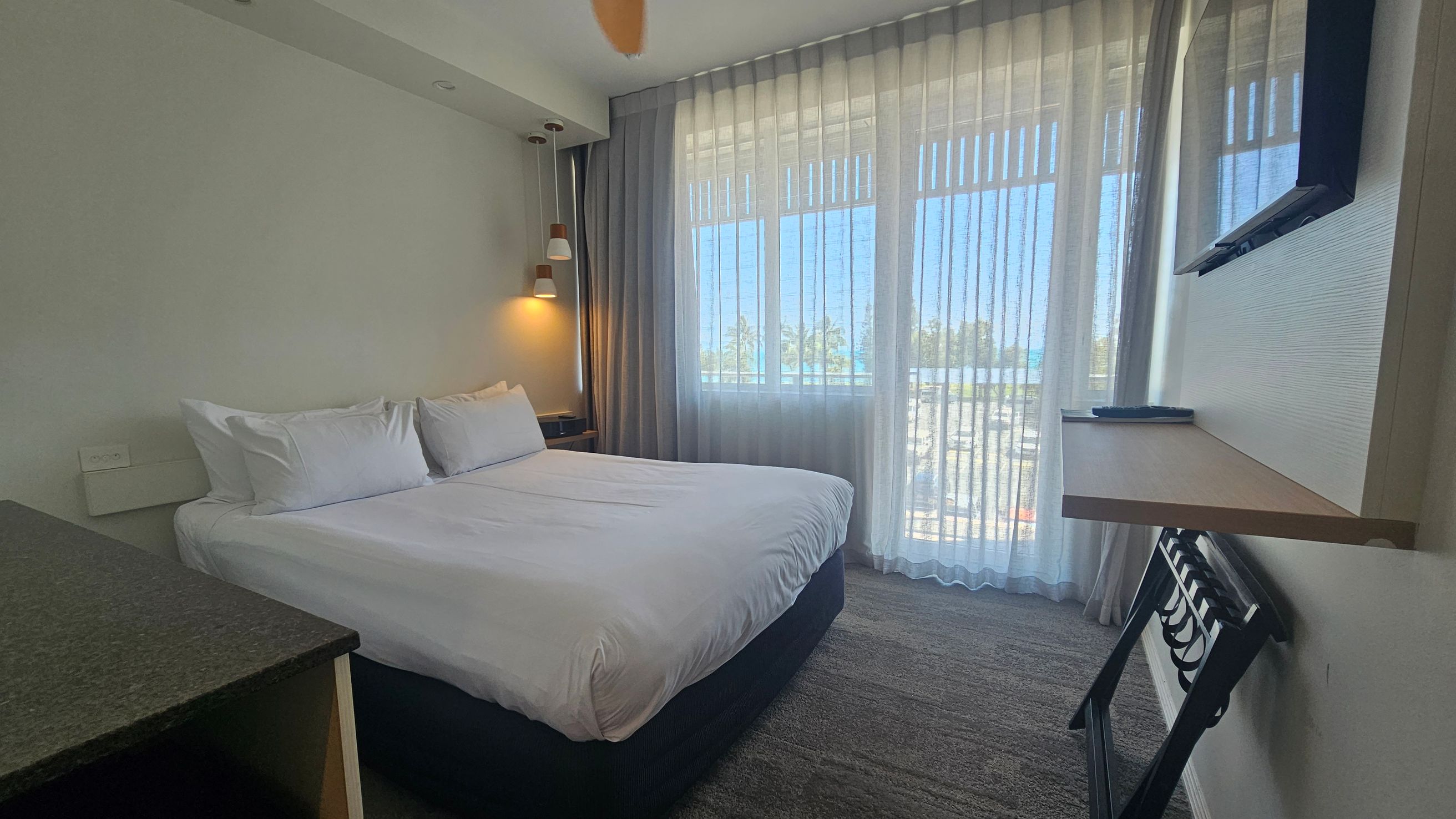 A hotel room with a large bed on the left, covered in white linens and pillows. Above the bed, a wall lamp provides light. On the right, a flat-screen TV is mounted on the wall above a small shelf. Below the shelf is a luggage rack. The room has a large window with sheer curtains, allowing natural light to filter in. The floor is carpeted in a neutral tone. A hotel room with a large bed on the left, covered in white linens and pillows. Above the bed, a wall lamp provides light. On the right, a flat-screen TV is mounted on the wall above a small shelf. Below the shelf is a luggage rack. The room has a large window with sheer curtains, allowing natural light to filter in. The floor is carpeted in a neutral tone.