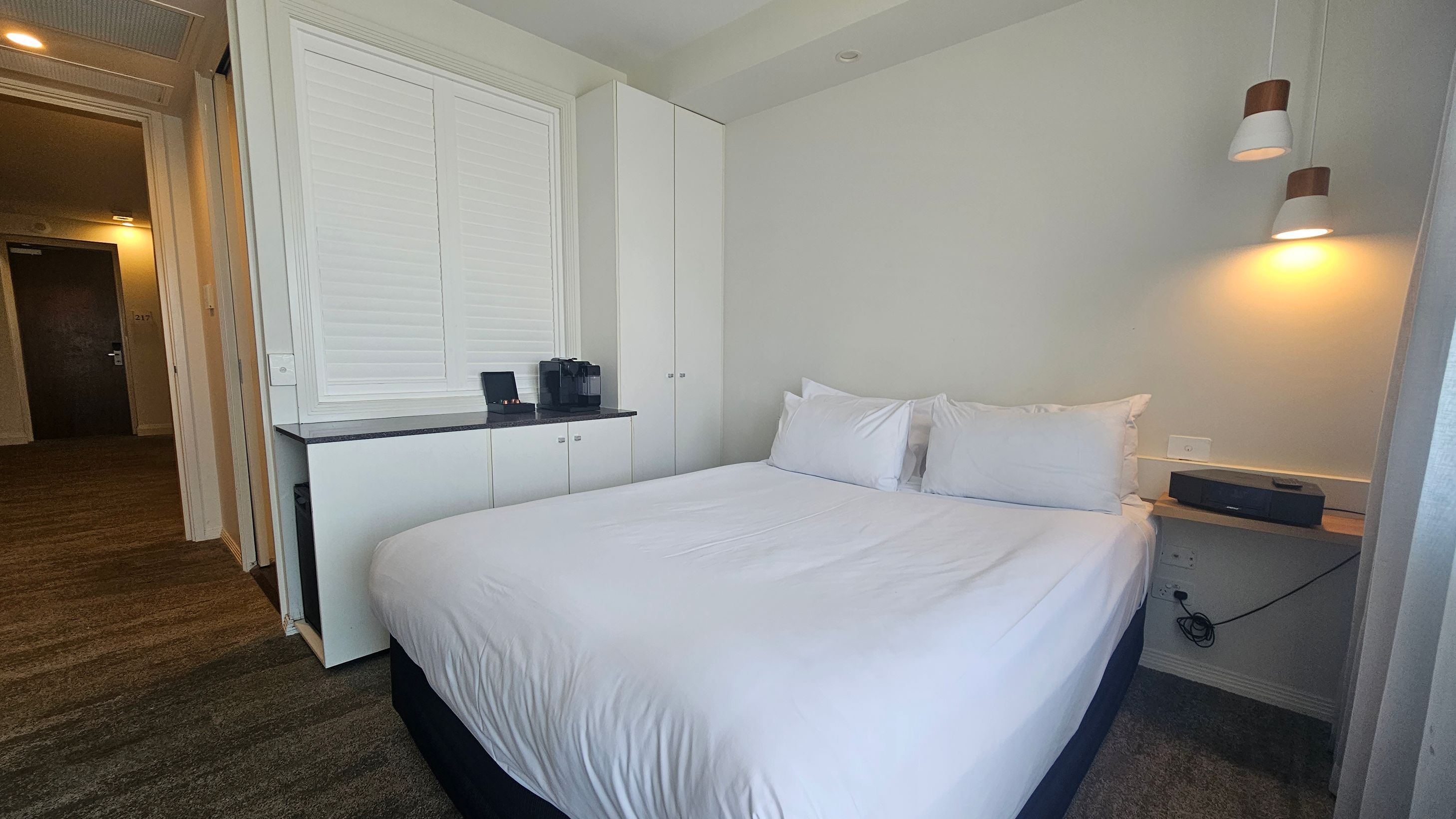 A hotel room with a neatly made double bed on the right, featuring white linens and two pillows. Above the bed, two pendant lights hang from the ceiling. On the left, there is a built-in cabinet with a countertop, holding a coffee machine and a tray. The floor is carpeted in a mix of green and brown tones. An open door leads to a hallway with a visible room number, 1217, on a dark door. The walls are light-coloured, and the ceiling is plain with recessed lighting. A hotel room with a neatly made double bed on the right, featuring white linens and two pillows. Above the bed, two pendant lights hang from the ceiling. On the left, there is a built-in cabinet with a countertop, holding a coffee machine and a tray. The floor is carpeted in a mix of green and brown tones. An open door leads to a hallway with a visible room number, 1217, on a dark door. The walls are light-coloured, and the ceiling is plain with recessed lighting.