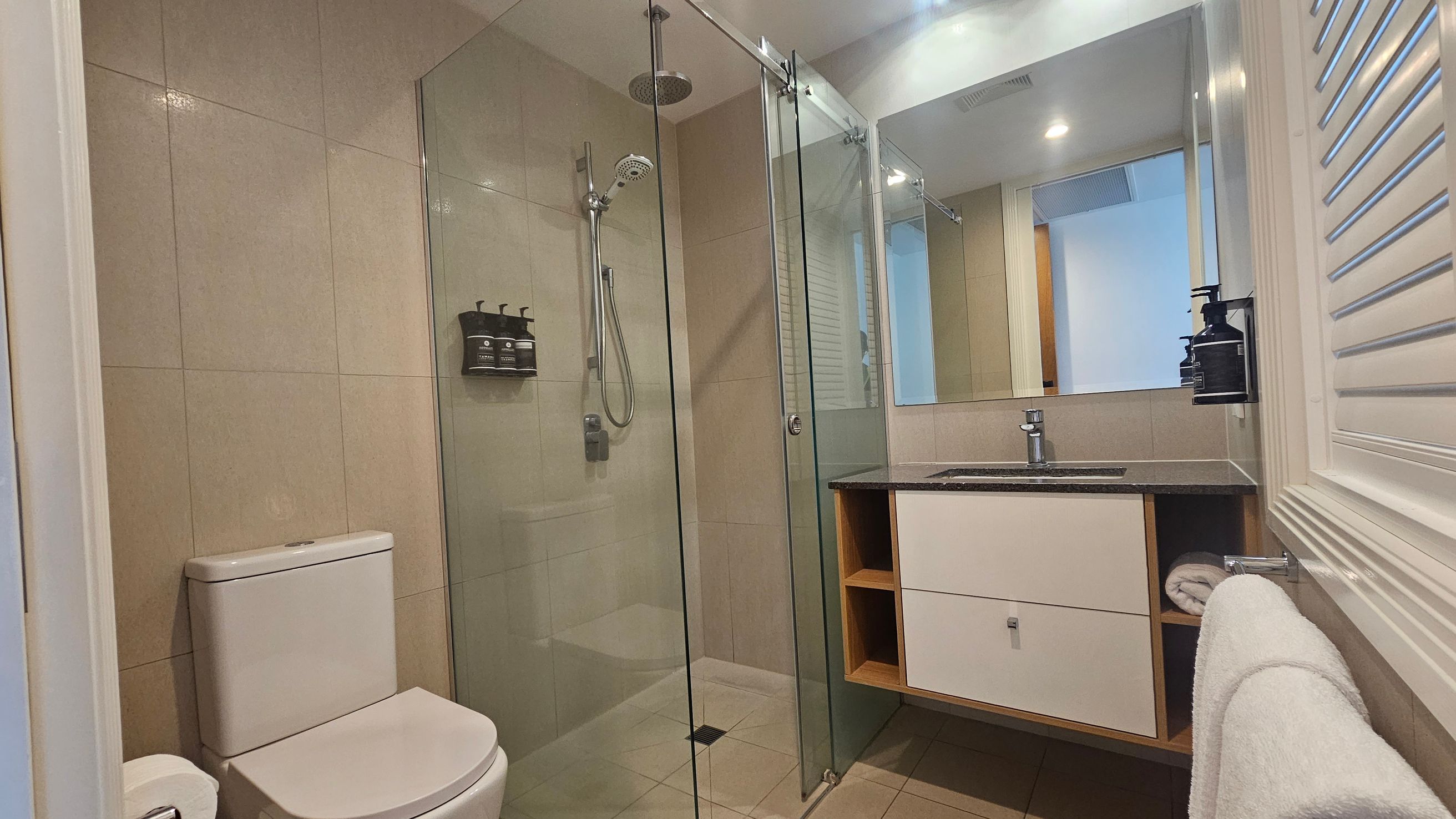 Modern bathroom with beige tiled walls and floor. On the left, a white toilet is positioned next to a glass-enclosed shower with a rainfall showerhead and mounted toiletries. On the right, a vanity with a black countertop holds a sink, with a large mirror above. Below the sink, there are open shelves and a towel rack with a white towel. The ceiling has recessed lighting, and a louvred panel is visible on the right wall. Modern bathroom with beige tiled walls and floor. On the left, a white toilet is positioned next to a glass-enclosed shower with a rainfall showerhead and mounted toiletries. On the right, a vanity with a black countertop holds a sink, with a large mirror above. Below the sink, there are open shelves and a towel rack with a white towel. The ceiling has recessed lighting, and a louvred panel is visible on the right wall.
