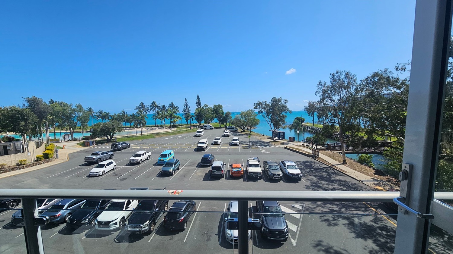 A view from a balcony overlooking a car park with several vehicles parked in rows. Beyond the car park, there are palm trees and a grassy area leading to a turquoise sea. On the right, a small bridge crosses over a narrow waterway. The scene is framed by a glass railing in the foreground. A view from a balcony overlooking a car park with several vehicles parked in rows. Beyond the car park, there are palm trees and a grassy area leading to a turquoise sea. On the right, a small bridge crosses over a narrow waterway. The scene is framed by a glass railing in the foreground.