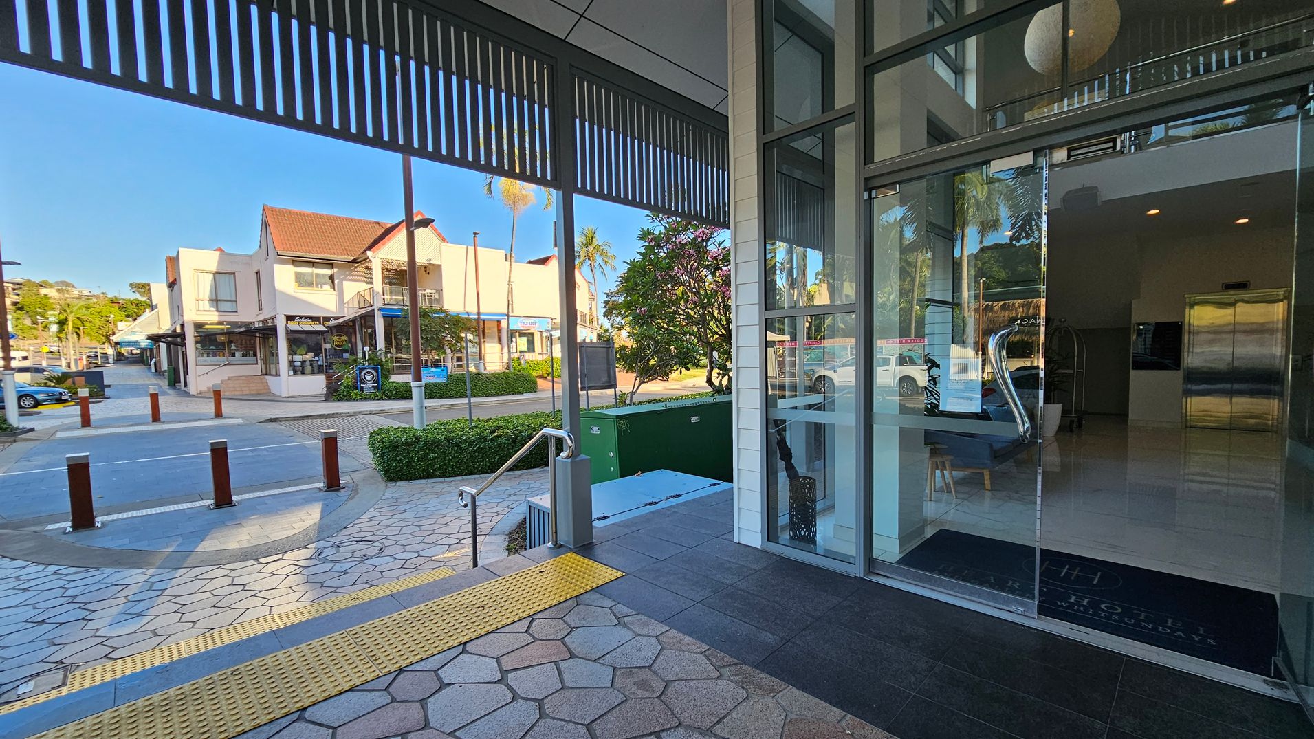 A modern building entrance with glass doors on the right, leading to a lobby with a shiny floor and an elevator. Outside, a paved area with hexagonal tiles and a yellow tactile strip. A small set of stairs with a metal railing descends to the left. Across the street, there are shops with red-tiled roofs and a few parked cars. Greenery, including a bush and a tree, is visible near the entrance. A modern building entrance with glass doors on the right, leading to a lobby with a shiny floor and an elevator. Outside, a paved area with hexagonal tiles and a yellow tactile strip. A small set of stairs with a metal railing descends to the left. Across the street, there are shops with red-tiled roofs and a few parked cars. Greenery, including a bush and a tree, is visible near the entrance.