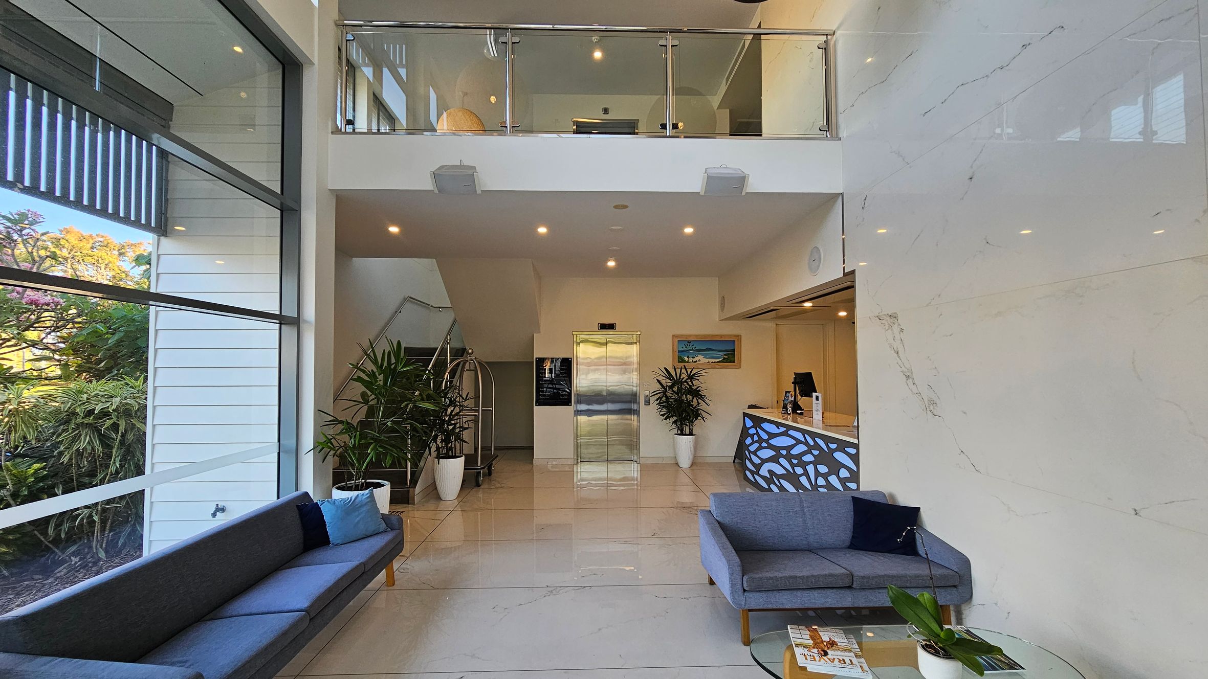 A modern lobby with a high ceiling and a mezzanine level. On the left, large windows reveal lush greenery outside. A long grey sofa with blue cushions is positioned against the window. In the centre, a lift is flanked by potted plants and a staircase. On the right, a reception desk with a decorative panel is accompanied by a grey sofa and a glass coffee table with magazines and a plant. The floor is polished and light-coloured, complementing the sleek, contemporary design. A modern lobby with a high ceiling and a mezzanine level. On the left, large windows reveal lush greenery outside. A long grey sofa with blue cushions is positioned against the window. In the centre, a lift is flanked by potted plants and a staircase. On the right, a reception desk with a decorative panel is accompanied by a grey sofa and a glass coffee table with magazines and a plant. The floor is polished and light-coloured, complementing the sleek, contemporary design.