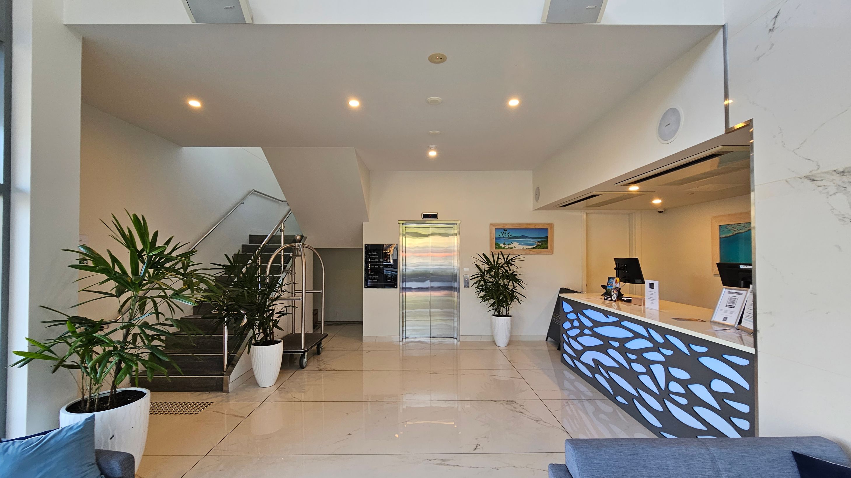 A modern lobby area with a staircase on the left, featuring metal railings and potted plants. In the centre, there's a lift with a metallic door, flanked by more plants and a wall-mounted painting. On the right, a reception desk with a decorative front panel, two computer monitors, and informational materials. The floor is tiled in a light colour, and the ceiling has recessed lighting. A modern lobby area with a staircase on the left, featuring metal railings and potted plants. In the centre, there's a lift with a metallic door, flanked by more plants and a wall-mounted painting. On the right, a reception desk with a decorative front panel, two computer monitors, and informational materials. The floor is tiled in a light colour, and the ceiling has recessed lighting.