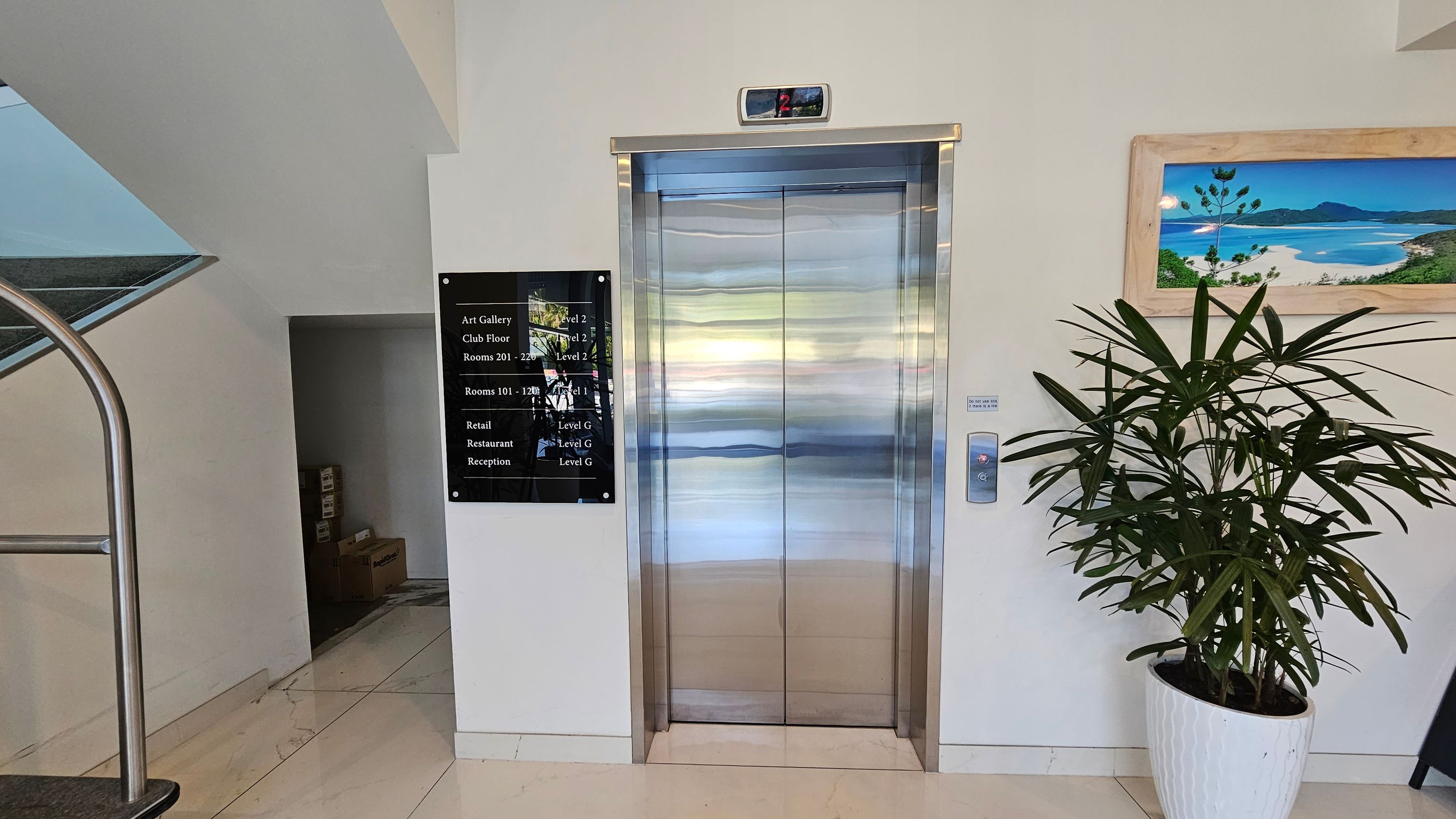 A modern lift with shiny metallic doors is centred on a light-coloured wall. Above the lift, a digital display shows the number "2". To the left, a black directory sign lists various locations and levels. A staircase with a metal railing is partially visible. On the right, a potted plant with long, slender leaves stands next to a framed landscape picture. The floor is tiled in a light colour. A modern lift with shiny metallic doors is centred on a light-coloured wall. Above the lift, a digital display shows the number "2". To the left, a black directory sign lists various locations and levels. A staircase with a metal railing is partially visible. On the right, a potted plant with long, slender leaves stands next to a framed landscape picture. The floor is tiled in a light colour.