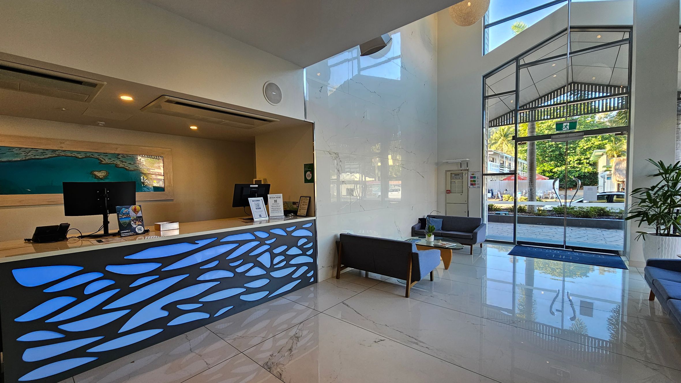 A modern reception area with a sleek desk on the left, featuring a blue abstract design and two computer monitors. Behind the desk is a large framed image of a coastal scene. On the right, there are glass doors leading outside, with a view of greenery and buildings. Two grey sofas and a wooden coffee table are positioned near the entrance. The floor is polished marble, and the ceiling is high with recessed lighting. A modern reception area with a sleek desk on the left, featuring a blue abstract design and two computer monitors. Behind the desk is a large framed image of a coastal scene. On the right, there are glass doors leading outside, with a view of greenery and buildings. Two grey sofas and a wooden coffee table are positioned near the entrance. The floor is polished marble, and the ceiling is high with recessed lighting.