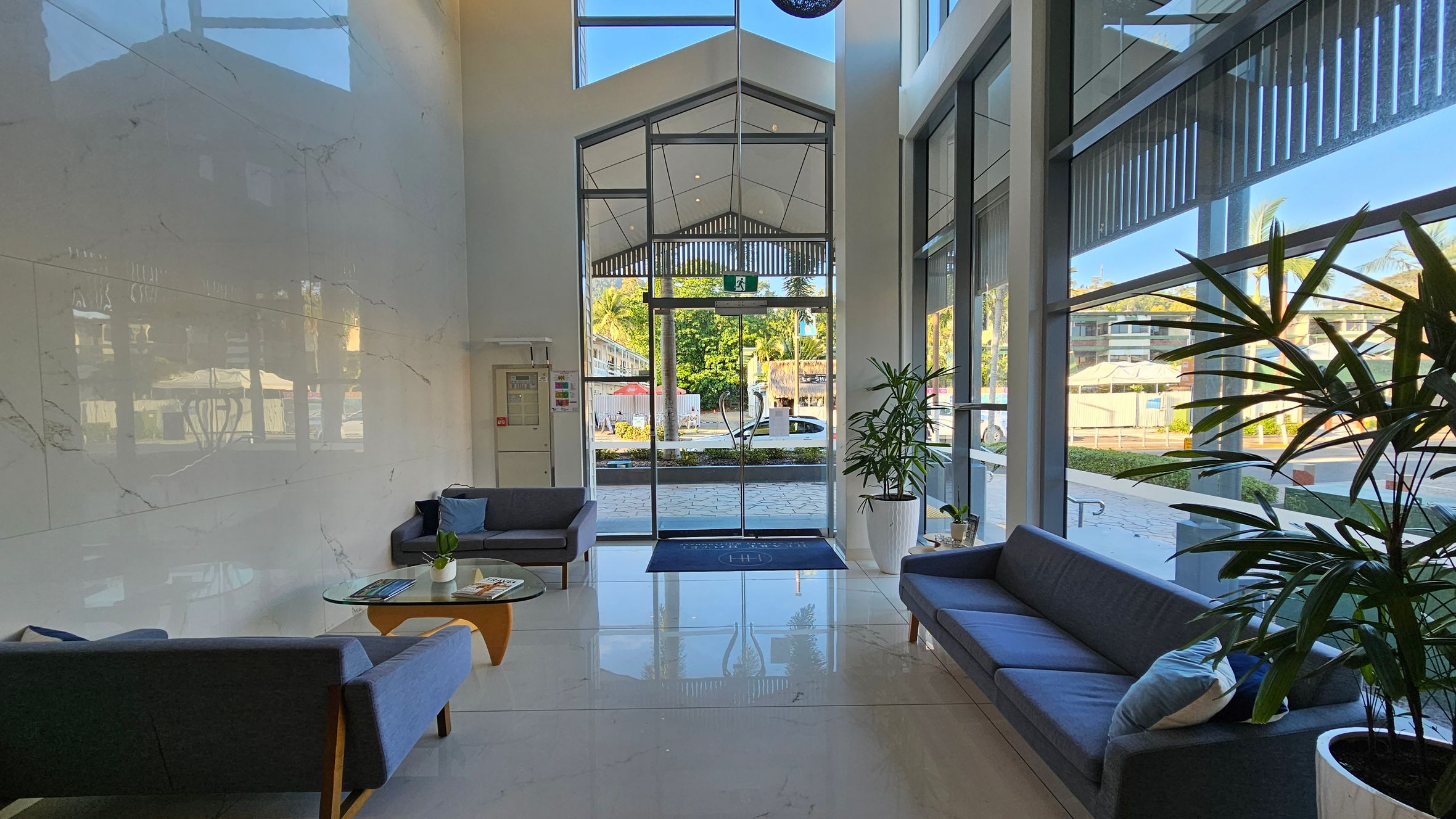 A modern lobby with a high ceiling and large glass entrance doors. On the left, there are two grey sofas with blue cushions and a glass coffee table with magazines and a small plant. On the right, another grey sofa with a cushion is next to a large potted plant. The floor is shiny and tiled, reflecting the furniture and natural light. Outside, greenery and a street are visible through the glass. A modern lobby with a high ceiling and large glass entrance doors. On the left, there are two grey sofas with blue cushions and a glass coffee table with magazines and a small plant. On the right, another grey sofa with a cushion is next to a large potted plant. The floor is shiny and tiled, reflecting the furniture and natural light. Outside, greenery and a street are visible through the glass.
