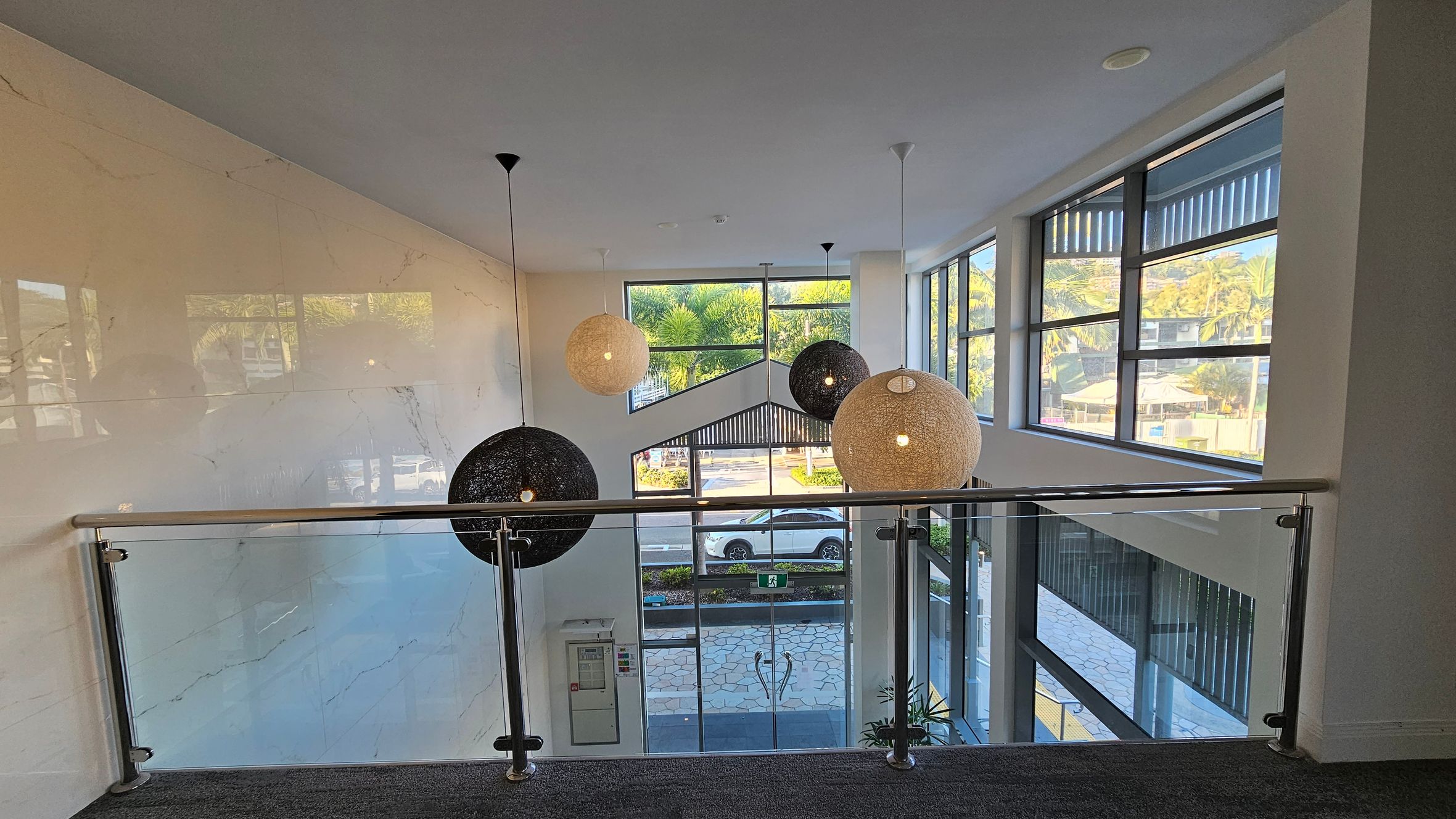 A modern interior with large windows on the right, allowing natural light to illuminate the space. In the centre, three spherical pendant lights hang from the ceiling, two black and one beige. A glass railing with metal supports runs across the foreground. The floor is carpeted in a dark grey tone. Outside, a view of a street with a parked car and greenery is visible through the windows. A modern interior with large windows on the right, allowing natural light to illuminate the space. In the centre, three spherical pendant lights hang from the ceiling, two black and one beige. A glass railing with metal supports runs across the foreground. The floor is carpeted in a dark grey tone. Outside, a view of a street with a parked car and greenery is visible through the windows.