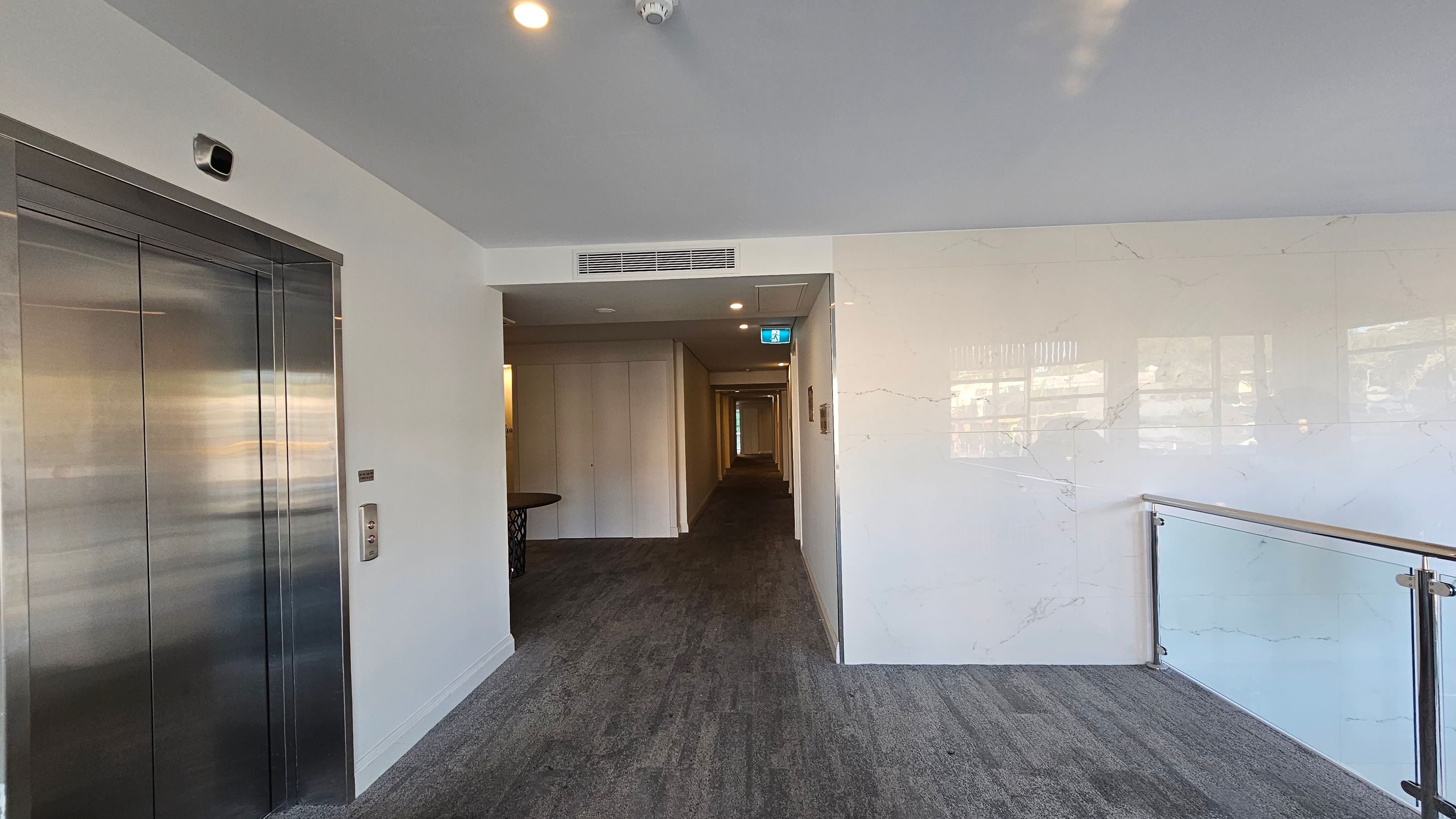 On the left, there is a metallic lift door with a control panel beside it. In the centre, a hallway extends into the distance, with a table visible near the entrance. The right side features a glass railing with a wooden handrail, overlooking a lower level. The floor is carpeted in a grey tone, and the ceiling is white with recessed lighting and a smoke detector. On the left, there is a metallic lift door with a control panel beside it. In the centre, a hallway extends into the distance, with a table visible near the entrance. The right side features a glass railing with a wooden handrail, overlooking a lower level. The floor is carpeted in a grey tone, and the ceiling is white with recessed lighting and a smoke detector.