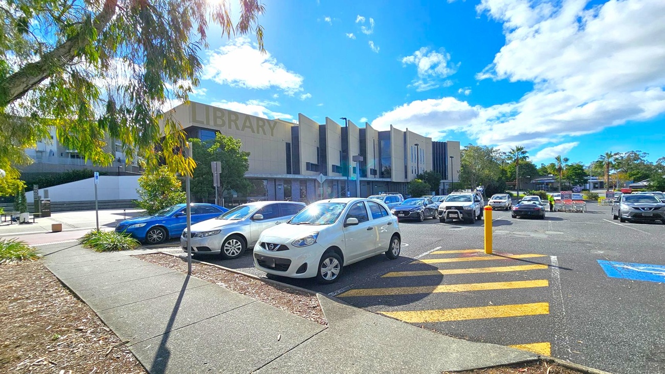 A car park with several vehicles, including a white car in the foreground. On the left, a tree with green leaves partially shades the area. The background features a large building with the word "LIBRARY" visible on its facade. The sky is blue with scattered clouds. A yellow-striped pedestrian crossing and a blue disabled parking space are visible on the asphalt. A car park with several vehicles, including a white car in the foreground. On the left, a tree with green leaves partially shades the area. The background features a large building with the word "LIBRARY" visible on its facade. The sky is blue with scattered clouds. A yellow-striped pedestrian crossing and a blue disabled parking space are visible on the asphalt.