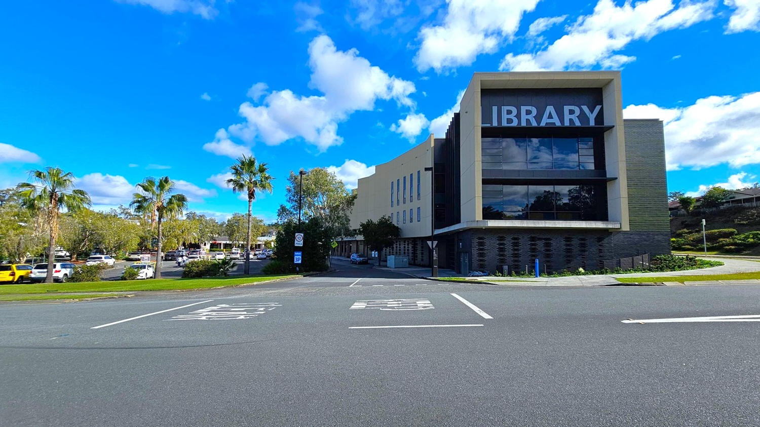 A modern library building is on the right, with large windows and the word "LIBRARY" prominently displayed. The foreground features a road with marked parking spaces. On the left, there are several palm trees and a parking area filled with cars. The background includes more trees and a clear blue sky with scattered clouds. A modern library building is on the right, with large windows and the word "LIBRARY" prominently displayed. The foreground features a road with marked parking spaces. On the left, there are several palm trees and a parking area filled with cars. The background includes more trees and a clear blue sky with scattered clouds.