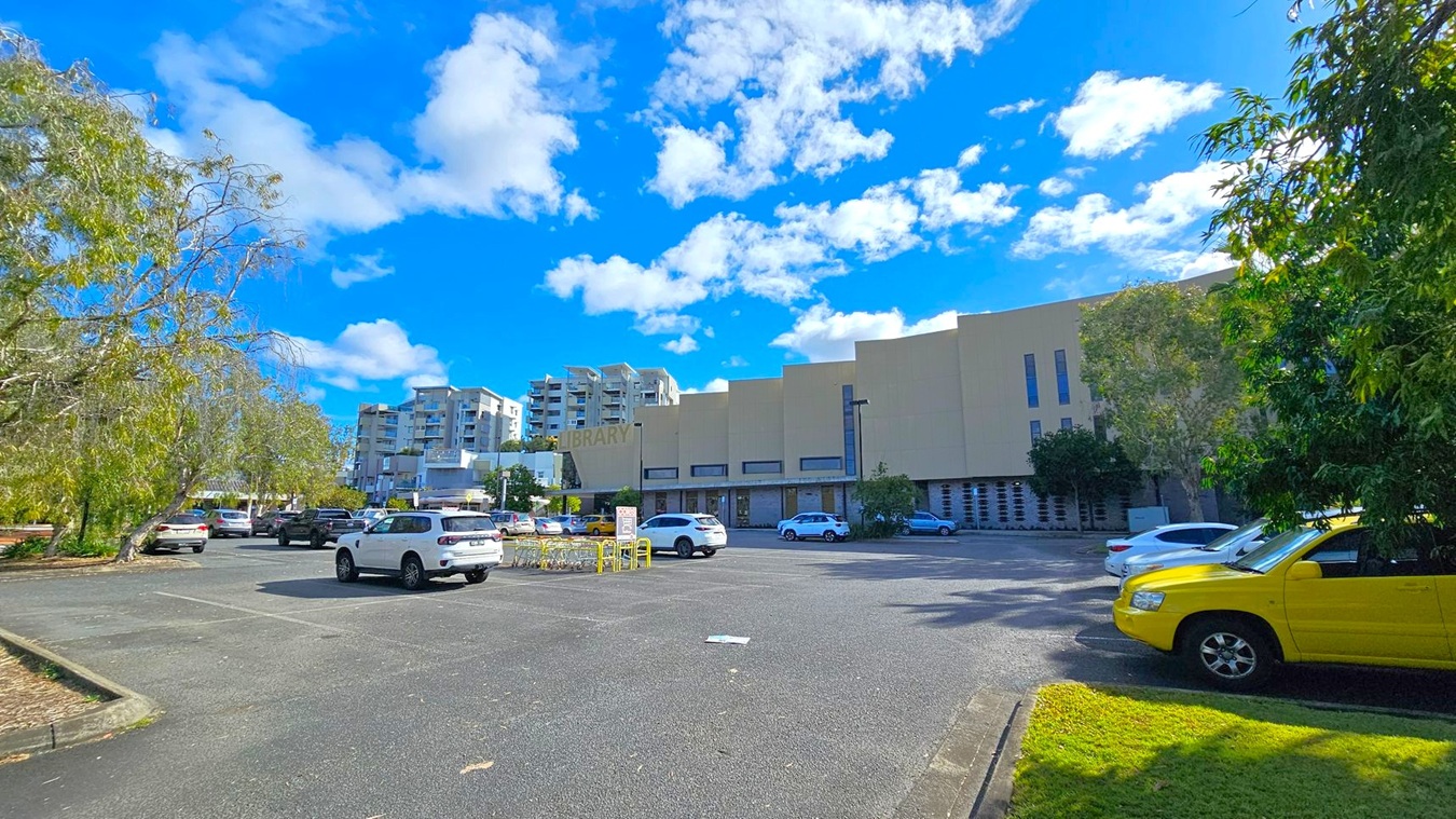 A car park with several vehicles, including a prominent yellow car on the right. In the background, a large beige building with the word "LIBRARY" is visible. To the left, there are trees with sparse foliage, and several multi-storey buildings can be seen in the distance. The ground is paved, and there is a small patch of grass near the yellow car. A car park with several vehicles, including a prominent yellow car on the right. In the background, a large beige building with the word "LIBRARY" is visible. To the left, there are trees with sparse foliage, and several multi-storey buildings can be seen in the distance. The ground is paved, and there is a small patch of grass near the yellow car.