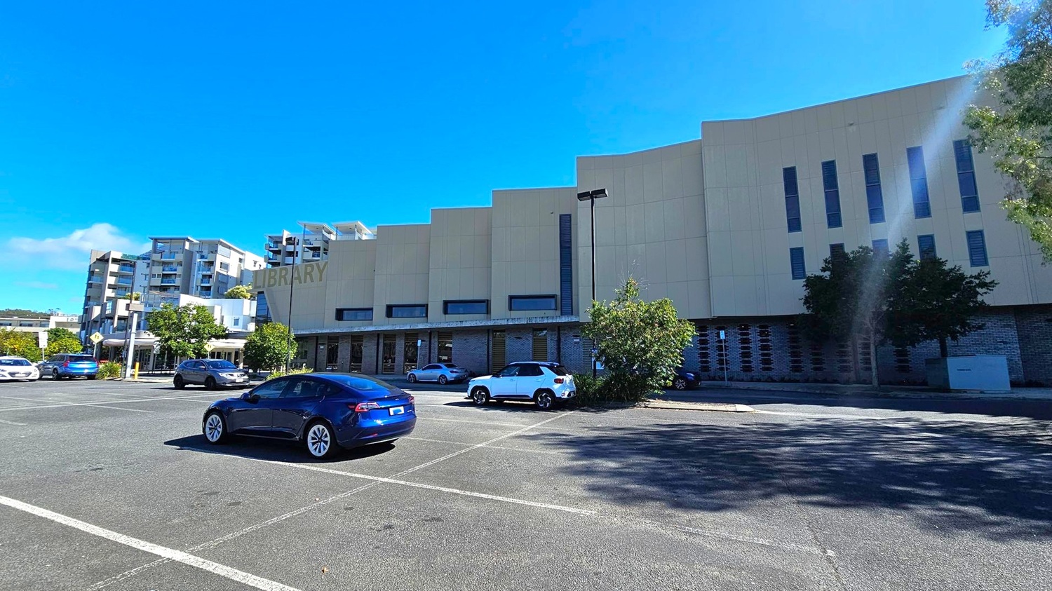 A large, modern building with the word "LIBRARY" on its facade is on the right. The structure has a series of vertical windows and a light-coloured exterior. In the foreground, a car park with several vehicles, including a blue car and a white SUV. On the left, there are multi-storey residential buildings with balconies. Trees and shrubs are scattered around the area, providing some greenery. The ground is paved with marked parking spaces. A large, modern building with the word "LIBRARY" on its facade is on the right. The structure has a series of vertical windows and a light-coloured exterior. In the foreground, a car park with several vehicles, including a blue car and a white SUV. On the left, there are multi-storey residential buildings with balconies. Trees and shrubs are scattered around the area, providing some greenery. The ground is paved with marked parking spaces.