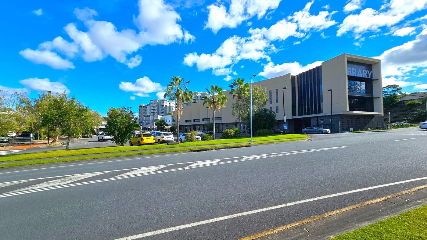 A modern building with the word "BRARY" visible on the right side, likely part of a library sign. The structure is surrounded by palm trees and other greenery. Several cars are parked in a lot to the left, including a yellow one. The foreground features a multi-lane road with white markings. In the background, there are additional buildings and more trees under a blue sky with scattered clouds. A modern building with the word "BRARY" visible on the right side, likely part of a library sign. The structure is surrounded by palm trees and other greenery. Several cars are parked in a lot to the left, including a yellow one. The foreground features a multi-lane road with white markings. In the background, there are additional buildings and more trees under a blue sky with scattered clouds.