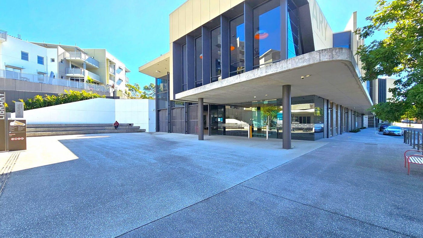 On the left, there is a modern building with white walls and balconies, surrounded by greenery. A person is sitting on steps near a sign that reads "Library" and "Cultural". In the centre, a large building with tall glass windows and a protruding roof supported by pillars. On the right, a tree provides shade over a parked car and a red bench. The ground is paved with a textured surface, and the area is well-lit by natural light. On the left, there is a modern building with white walls and balconies, surrounded by greenery. A person is sitting on steps near a sign that reads "Library" and "Cultural". In the centre, a large building with tall glass windows and a protruding roof supported by pillars. On the right, a tree provides shade over a parked car and a red bench. The ground is paved with a textured surface, and the area is well-lit by natural light.