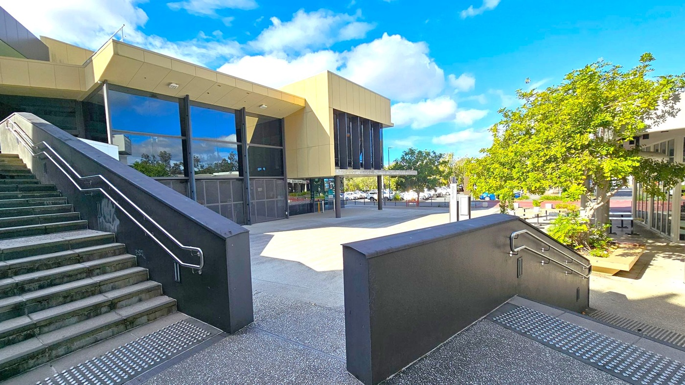 A modern building with large glass windows and beige cladding is in the centre. On the left, a wide staircase with metal railings leads up to the entrance. On the right, a tree with green foliage provides shade over a paved area. In the background, a car park is visible with several parked cars. The ground is a mix of textured and smooth concrete surfaces. A modern building with large glass windows and beige cladding is in the centre. On the left, a wide staircase with metal railings leads up to the entrance. On the right, a tree with green foliage provides shade over a paved area. In the background, a car park is visible with several parked cars. The ground is a mix of textured and smooth concrete surfaces.