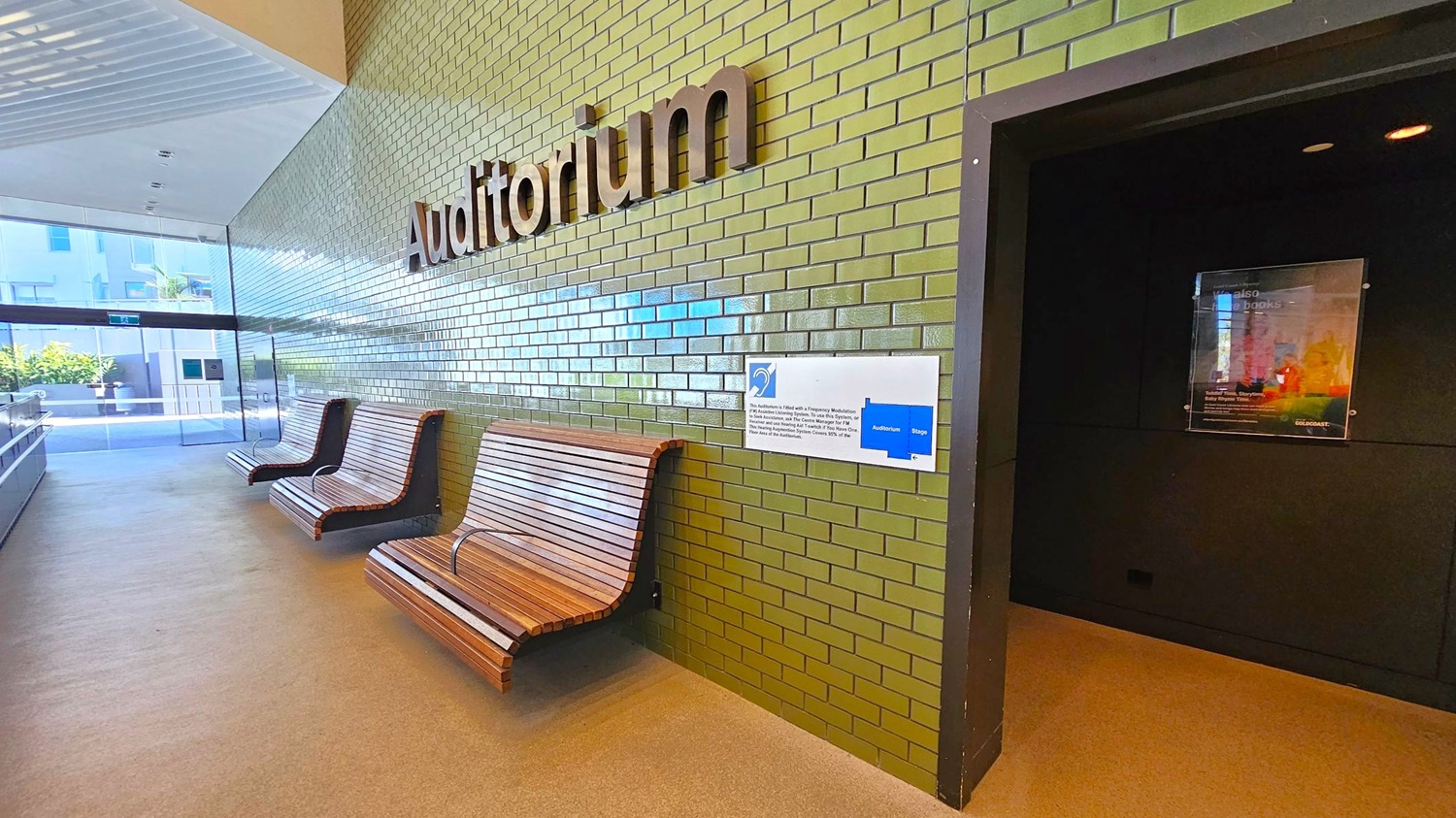 A hallway with green tiled walls and a sign reading "Auditorium" in large letters. On the left, there are three wooden benches with curved designs. A glass door at the end of the hallway reveals an outdoor area with plants. On the right, a dark entrance leads into another room, with a poster displayed on the wall. The floor is a light-coloured, smooth surface, and the ceiling is white with recessed lighting. A hallway with green tiled walls and a sign reading "Auditorium" in large letters. On the left, there are three wooden benches with curved designs. A glass door at the end of the hallway reveals an outdoor area with plants. On the right, a dark entrance leads into another room, with a poster displayed on the wall. The floor is a light-coloured, smooth surface, and the ceiling is white with recessed lighting.