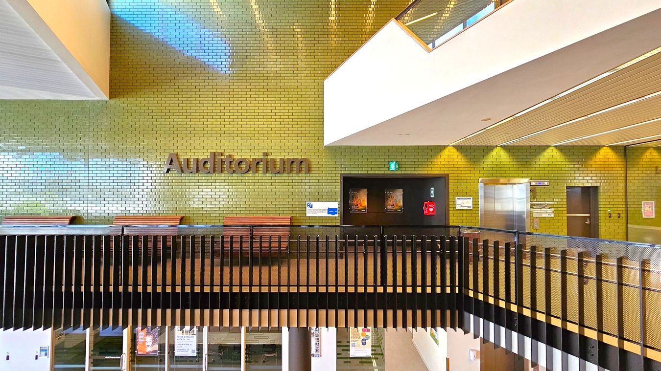 A modern interior with a green-tiled wall featuring the word "Auditorium" in large letters. Below, there are wooden benches and a black railing. On the right, a lift with metallic doors and a dark doorway are visible. The ceiling has wooden panels with embedded lights. The floor is not visible, but the lower level can be seen through the railing, showing glass doors and posters. A modern interior with a green-tiled wall featuring the word "Auditorium" in large letters. Below, there are wooden benches and a black railing. On the right, a lift with metallic doors and a dark doorway are visible. The ceiling has wooden panels with embedded lights. The floor is not visible, but the lower level can be seen through the railing, showing glass doors and posters.
