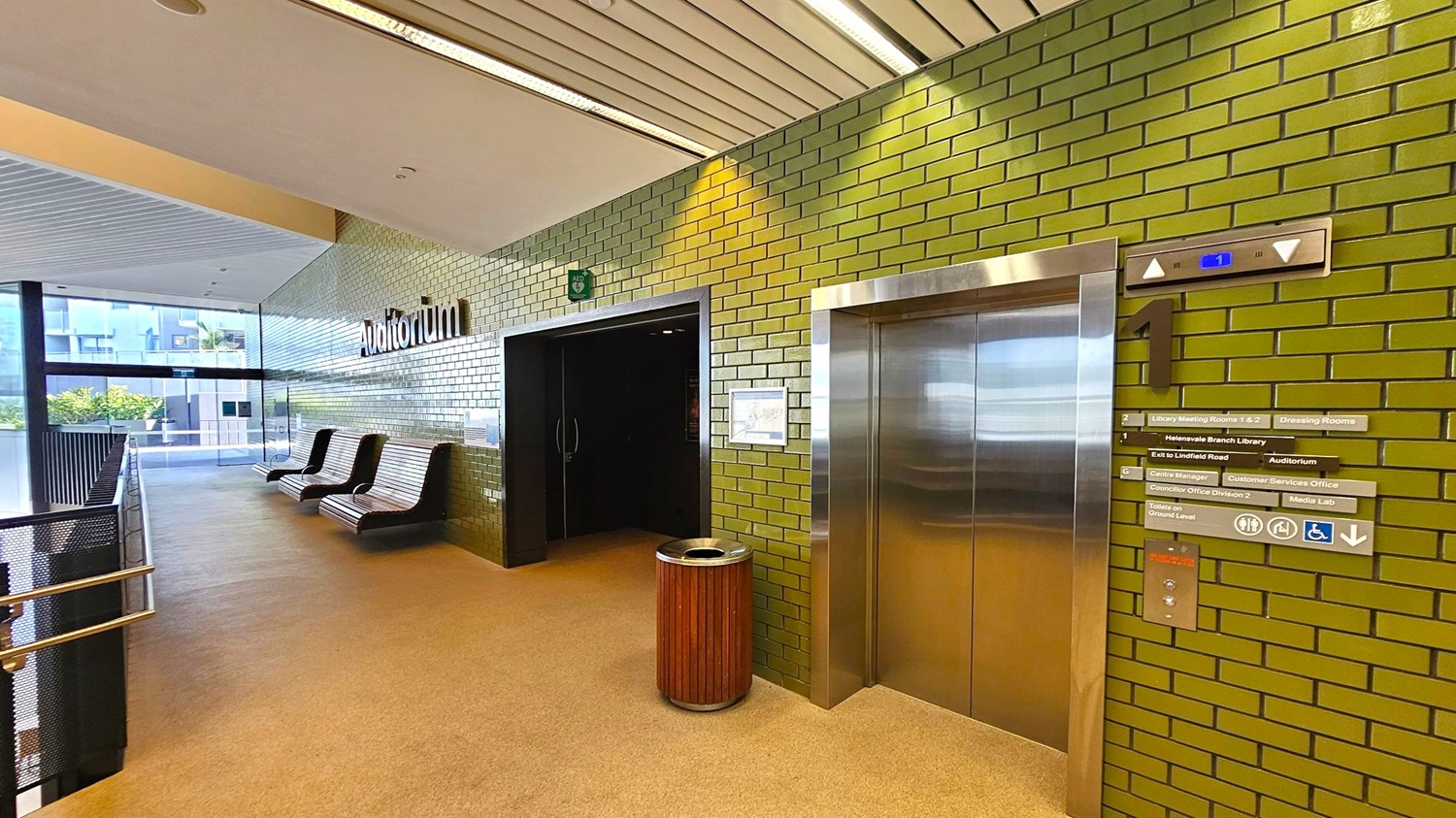A hallway with green brick walls and a beige carpeted floor. On the right, there's a stainless steel lift with floor indicators and directional signs. A wooden rubbish bin is placed nearby. The left side features a row of modern, curved benches against the wall, leading to a glass window with a view of greenery outside. The ceiling is lined with white panels and fluorescent lights. An entrance to an auditorium is visible next to the lift. A hallway with green brick walls and a beige carpeted floor. On the right, there's a stainless steel lift with floor indicators and directional signs. A wooden rubbish bin is placed nearby. The left side features a row of modern, curved benches against the wall, leading to a glass window with a view of greenery outside. The ceiling is lined with white panels and fluorescent lights. An entrance to an auditorium is visible next to the lift.