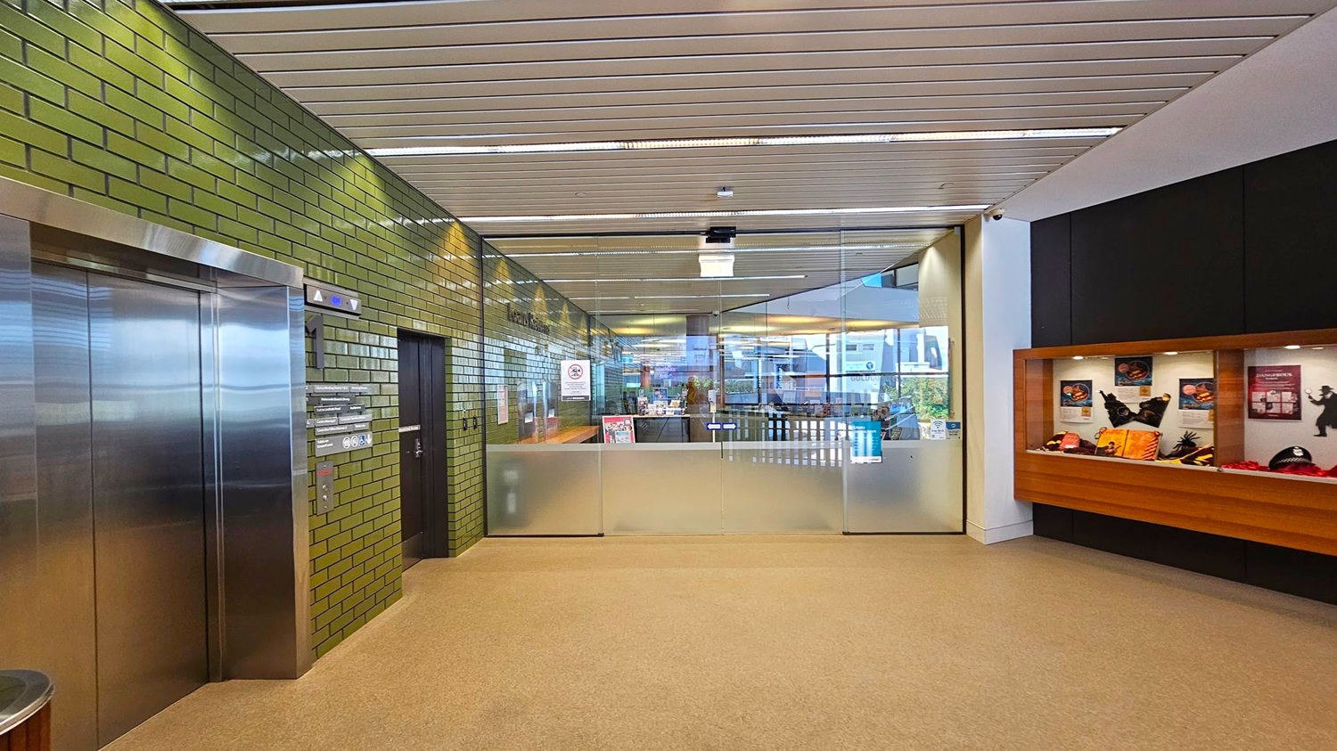 A hallway with a green tiled wall on the left featuring a stainless steel lift and a black door. The ceiling has a slatted design. In the centre, there's a glass partition with a frosted lower section, leading to another area. On the right, a display case with various items and posters is mounted on a wooden panel. The floor is carpeted in a light brown colour. A hallway with a green tiled wall on the left featuring a stainless steel lift and a black door. The ceiling has a slatted design. In the centre, there's a glass partition with a frosted lower section, leading to another area. On the right, a display case with various items and posters is mounted on a wooden panel. The floor is carpeted in a light brown colour.