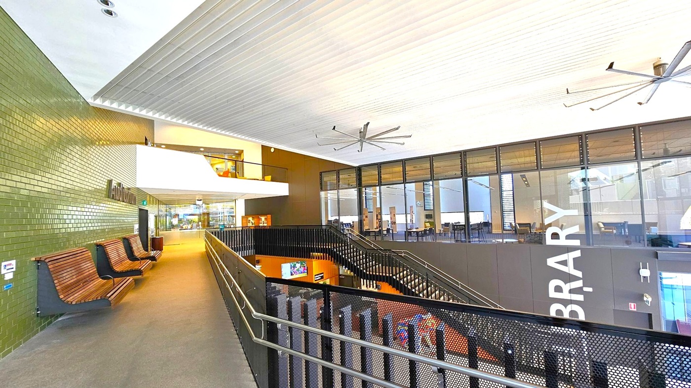 A modern interior with a high ceiling featuring large fans. On the left, there's a green-tiled wall with wooden benches and a sign for an auditorium. The right side has large glass windows with a view into a library area. A black metal railing lines the walkway, leading to a staircase descending to a lower level. The floor is carpeted, and the space is well-lit with natural and artificial light. A modern interior with a high ceiling featuring large fans. On the left, there's a green-tiled wall with wooden benches and a sign for an auditorium. The right side has large glass windows with a view into a library area. A black metal railing lines the walkway, leading to a staircase descending to a lower level. The floor is carpeted, and the space is well-lit with natural and artificial light.