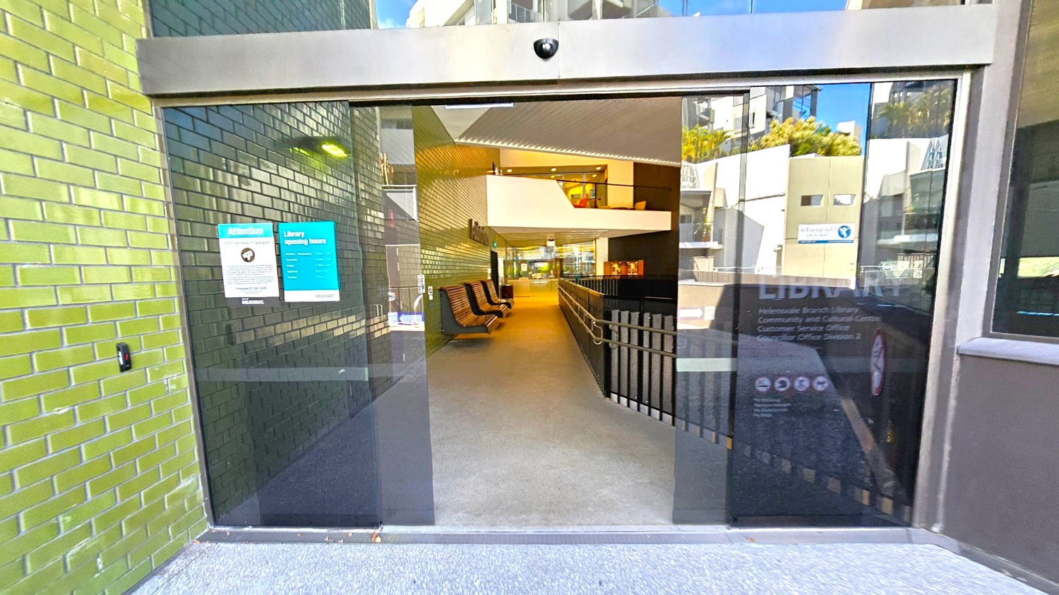 Glass entrance doors with signs about CCTV and library opening hours. The left wall is covered in green tiles. Inside, a corridor with wooden benches on the left and a railing on the right leads to a well-lit interior space. The floor is carpeted, and the ceiling has a modern design with recessed lighting. Reflections of nearby buildings are visible on the glass. Glass entrance doors with signs about CCTV and library opening hours. The left wall is covered in green tiles. Inside, a corridor with wooden benches on the left and a railing on the right leads to a well-lit interior space. The floor is carpeted, and the ceiling has a modern design with recessed lighting. Reflections of nearby buildings are visible on the glass.