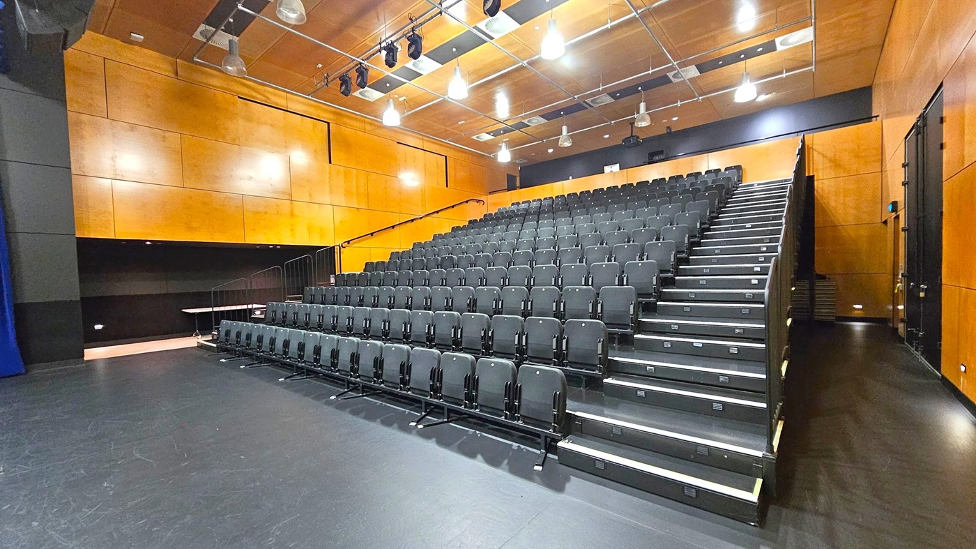A theatre with tiered seating on the right, featuring rows of black chairs. The floor is dark and smooth. The walls are clad in light wood panelling. The ceiling is also wooden, with exposed lighting fixtures and ventilation ducts. The left side has a small alcove with a table, and the right side has a narrow walkway. The space is well-lit with overhead lights. A theatre with tiered seating on the right, featuring rows of black chairs. The floor is dark and smooth. The walls are clad in light wood panelling. The ceiling is also wooden, with exposed lighting fixtures and ventilation ducts. The left side has a small alcove with a table, and the right side has a narrow walkway. The space is well-lit with overhead lights.