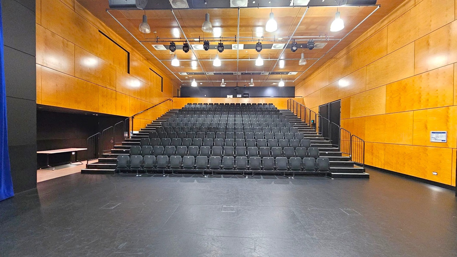 A theatre with tiered seating in the centre, featuring rows of black chairs. The walls are clad in light wood panelling. The ceiling is equipped with multiple hanging lights and stage equipment. On the left, a blue curtain is partially visible, while the right side has a wall-mounted sign. The floor is dark and smooth, contrasting with the light wood of the walls. A theatre with tiered seating in the centre, featuring rows of black chairs. The walls are clad in light wood panelling. The ceiling is equipped with multiple hanging lights and stage equipment. On the left, a blue curtain is partially visible, while the right side has a wall-mounted sign. The floor is dark and smooth, contrasting with the light wood of the walls.