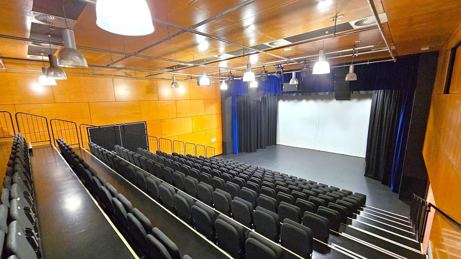 A modern auditorium with tiered seating on the left, featuring rows of black chairs. The stage is on the right, with a white screen and black curtains. The walls are wooden, and the ceiling is also wooden with exposed ductwork and several large, industrial-style pendant lights. The floor is dark, matching the seating area. A modern auditorium with tiered seating on the left, featuring rows of black chairs. The stage is on the right, with a white screen and black curtains. The walls are wooden, and the ceiling is also wooden with exposed ductwork and several large, industrial-style pendant lights. The floor is dark, matching the seating area.