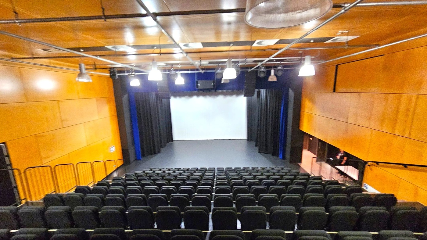 A theatre with rows of black seats facing a stage. The stage has a white backdrop and is flanked by black curtains. The walls are covered in light wood panelling. The ceiling is also wooden with exposed metal beams and several hanging lights. On the right, there is a small control booth with a person seated inside. The floor is dark, matching the seats. A theatre with rows of black seats facing a stage. The stage has a white backdrop and is flanked by black curtains. The walls are covered in light wood panelling. The ceiling is also wooden with exposed metal beams and several hanging lights. On the right, there is a small control booth with a person seated inside. The floor is dark, matching the seats.