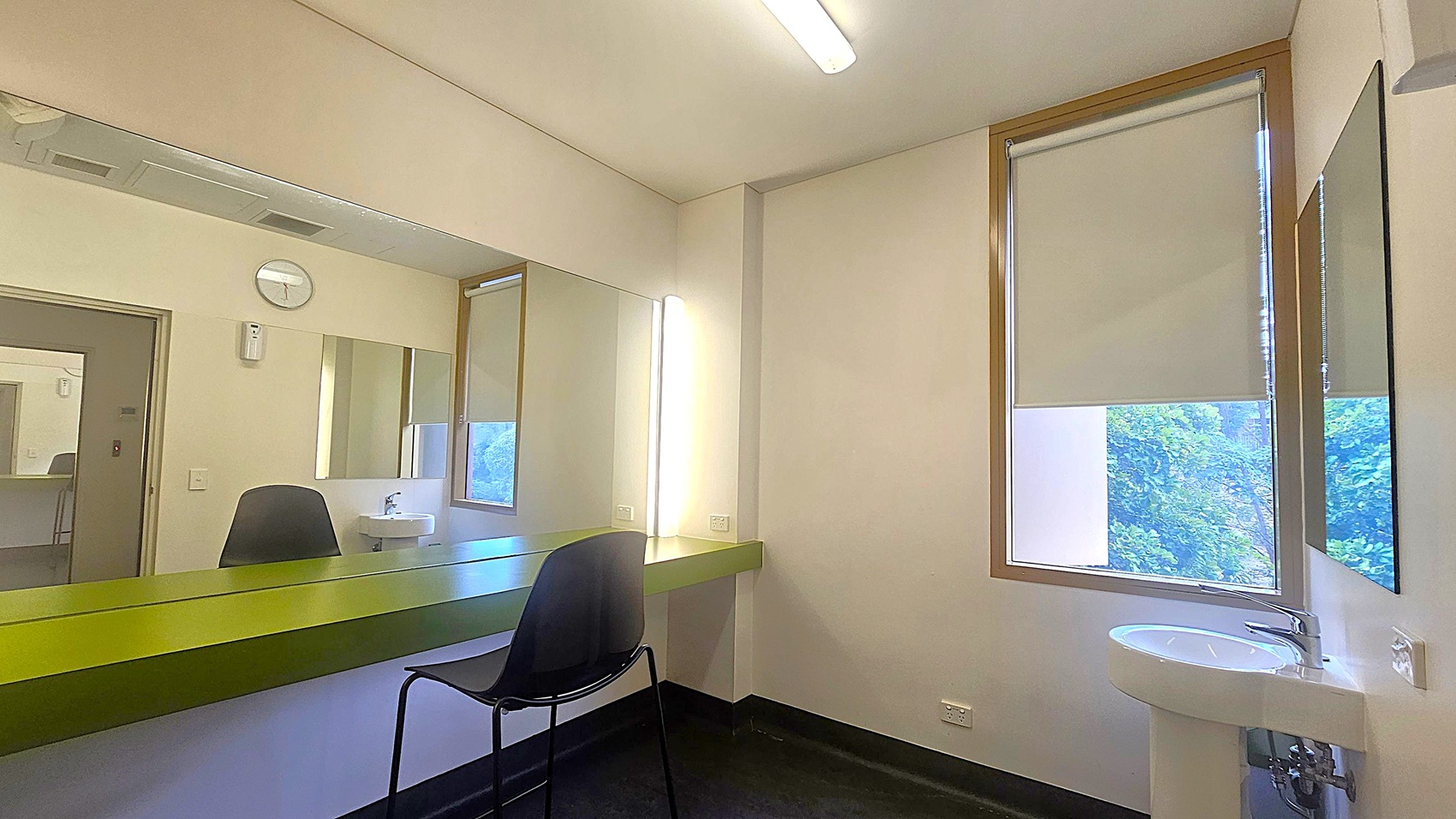 A room with a large mirror on the left wall, above a long green counter. A black chair is positioned in front of the counter. On the right, a window with a partially drawn blind reveals some greenery outside. Below the window is a small white sink. The ceiling has a fluorescent light fixture, and a clock is mounted on the wall above the mirror. The floor is dark, contrasting with the light-coloured walls. A room with a large mirror on the left wall, above a long green counter. A black chair is positioned in front of the counter. On the right, a window with a partially drawn blind reveals some greenery outside. Below the window is a small white sink. The ceiling has a fluorescent light fixture, and a clock is mounted on the wall above the mirror. The floor is dark, contrasting with the light-coloured walls.
