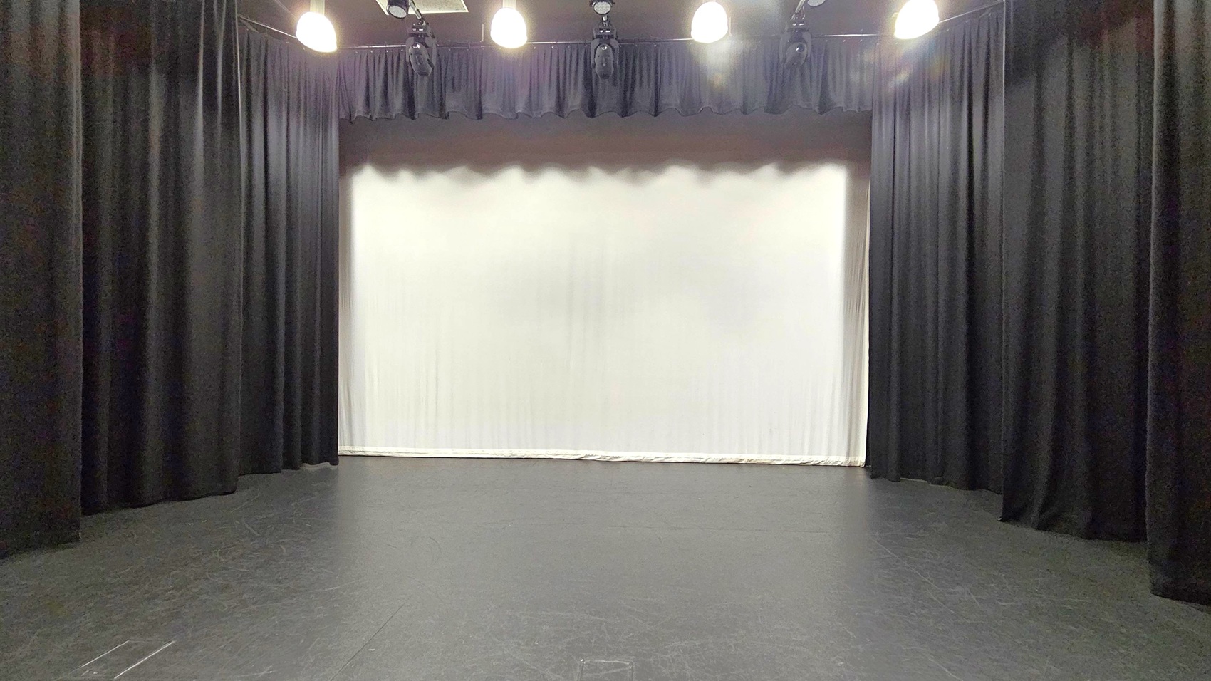 A stage with a black floor and black curtains on both sides. The background features a large white screen or backdrop. Overhead, several stage lights are mounted, casting light onto the stage area. The ceiling is dark, blending with the lighting equipment. A stage with a black floor and black curtains on both sides. The background features a large white screen or backdrop. Overhead, several stage lights are mounted, casting light onto the stage area. The ceiling is dark, blending with the lighting equipment.