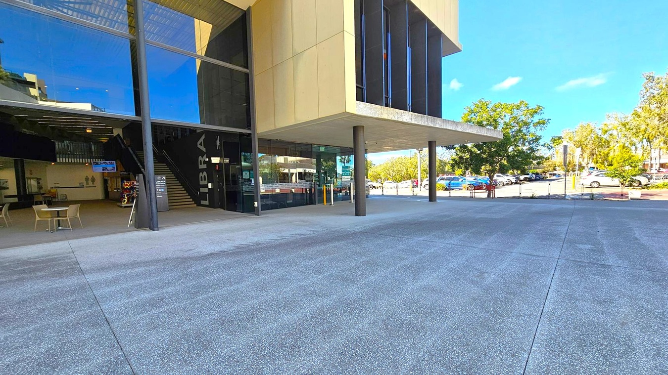 A modern building with large glass windows on the left, revealing an interior with tables, chairs, and a staircase. The word "LIBRA" is visible on the wall near the entrance. The building has a beige exterior with black columns supporting an overhang. On the right, there is a car park with several vehicles and trees in the background. The ground is paved with grey concrete. A modern building with large glass windows on the left, revealing an interior with tables, chairs, and a staircase. The word "LIBRA" is visible on the wall near the entrance. The building has a beige exterior with black columns supporting an overhang. On the right, there is a car park with several vehicles and trees in the background. The ground is paved with grey concrete.