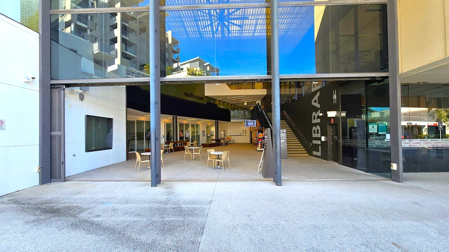 The image shows the entrance to a modern building with large glass windows. In the centre, there are tables and chairs arranged in an open area. To the right, a staircase leads upwards, with the word "LIBRARY" visible on the wall. The left side features a white wall with a dark window. The floor is a mix of concrete and tiled surfaces, and the ceiling has a grid-like structure with reflections of nearby buildings visible in the glass. The image shows the entrance to a modern building with large glass windows. In the centre, there are tables and chairs arranged in an open area. To the right, a staircase leads upwards, with the word "LIBRARY" visible on the wall. The left side features a white wall with a dark window. The floor is a mix of concrete and tiled surfaces, and the ceiling has a grid-like structure with reflections of nearby buildings visible in the glass.