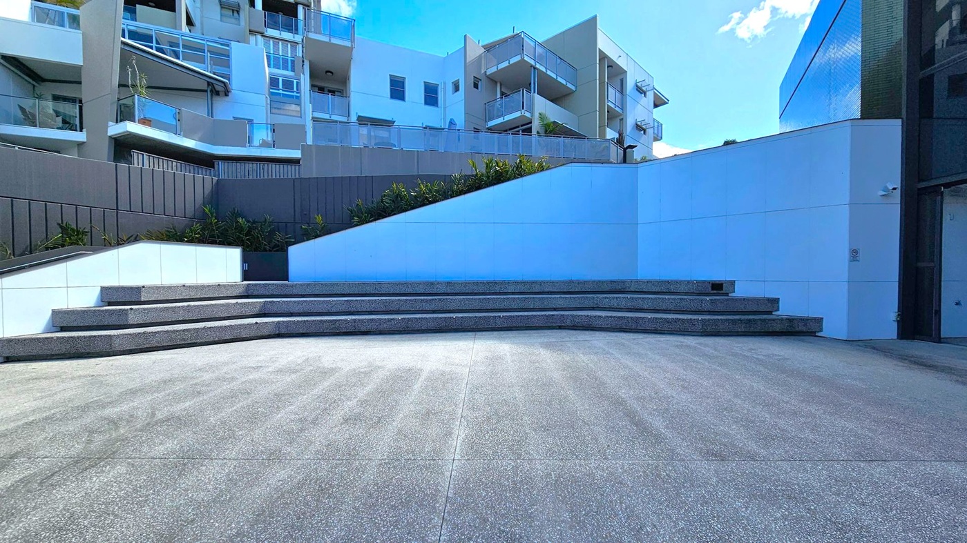 In the foreground, there is a wide, grey concrete area leading to a series of tiered steps. The steps are made of a darker material and are positioned centrally. In the background, modern apartment buildings with balconies and glass railings are visible. The left side features a dark panelled wall with some greenery at its base. The right side has a smooth, white wall with a slight curve. The sky is partially visible above the buildings. In the foreground, there is a wide, grey concrete area leading to a series of tiered steps. The steps are made of a darker material and are positioned centrally. In the background, modern apartment buildings with balconies and glass railings are visible. The left side features a dark panelled wall with some greenery at its base. The right side has a smooth, white wall with a slight curve. The sky is partially visible above the buildings.