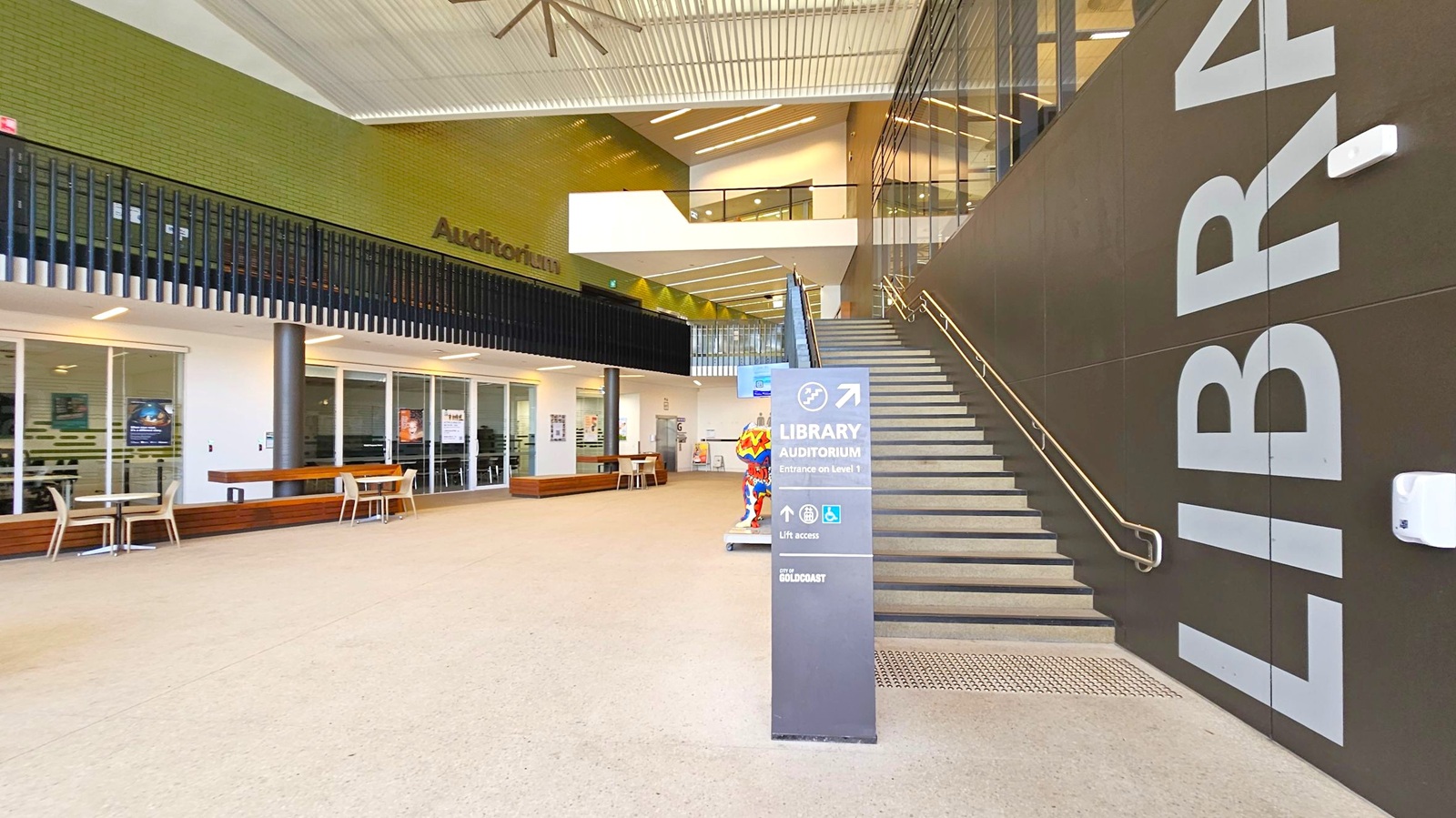 A modern interior space with a staircase on the right, featuring large letters spelling "LIBRARY" on the wall. A sign in front of the stairs indicates directions to the library and auditorium. On the left, there are glass doors with tables and chairs outside. Above, a green brick wall with the word "Auditorium" and a black railing. The ceiling has a large fan and linear lighting. The floor is light-coloured and smooth. A modern interior space with a staircase on the right, featuring large letters spelling "LIBRARY" on the wall. A sign in front of the stairs indicates directions to the library and auditorium. On the left, there are glass doors with tables and chairs outside. Above, a green brick wall with the word "Auditorium" and a black railing. The ceiling has a large fan and linear lighting. The floor is light-coloured and smooth.