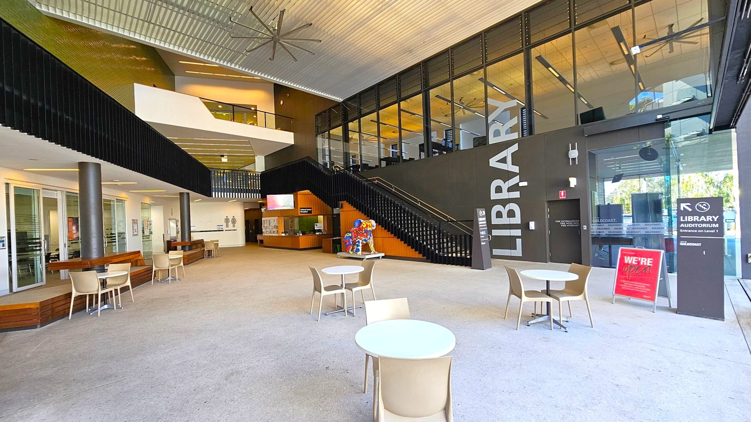 A spacious library entrance with a high ceiling and large fan. On the right, a black wall with "LIBRARY" in large letters and a glass entrance. A staircase leads to an upper level with glass panels. In the centre, a colourful sculpture is near the stairs. On the left, a seating area with tables and chairs, and a reception desk. The floor is light-coloured, and there are signs indicating the library is open. A spacious library entrance with a high ceiling and large fan. On the right, a black wall with "LIBRARY" in large letters and a glass entrance. A staircase leads to an upper level with glass panels. In the centre, a colourful sculpture is near the stairs. On the left, a seating area with tables and chairs, and a reception desk. The floor is light-coloured, and there are signs indicating the library is open.