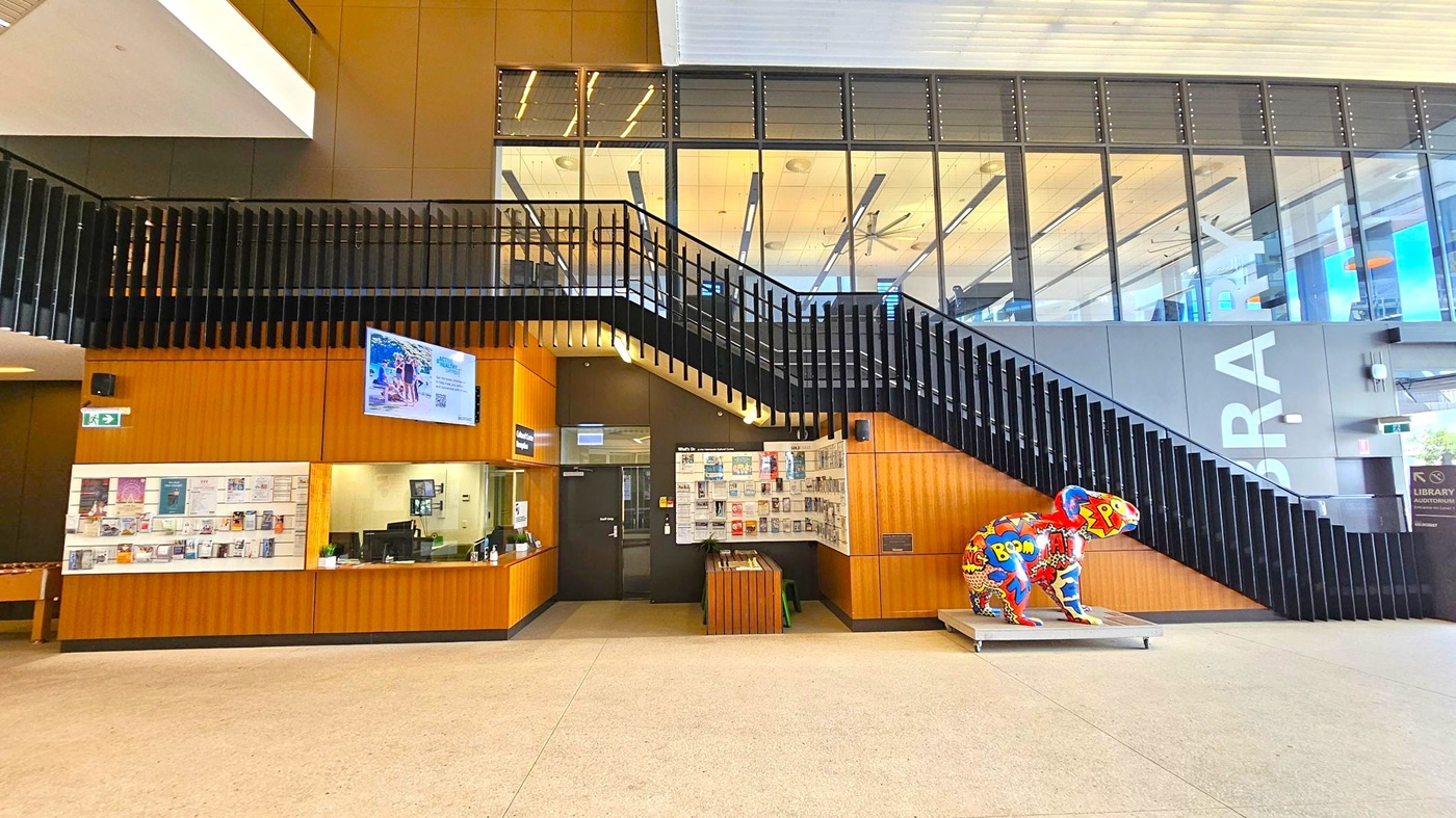 A modern interior with a staircase featuring black railings on the right. Below the stairs, a colourful sculpture of a koala with comic-style graphics is displayed. On the left, a wooden reception desk with a digital screen above it and a wall covered in posters and notices. The floor is light-coloured, and the ceiling has a grid pattern with visible lighting. Large windows above the staircase reveal an upper level with visible ceiling fans and lights. A modern interior with a staircase featuring black railings on the right. Below the stairs, a colourful sculpture of a koala with comic-style graphics is displayed. On the left, a wooden reception desk with a digital screen above it and a wall covered in posters and notices. The floor is light-coloured, and the ceiling has a grid pattern with visible lighting. Large windows above the staircase reveal an upper level with visible ceiling fans and lights.