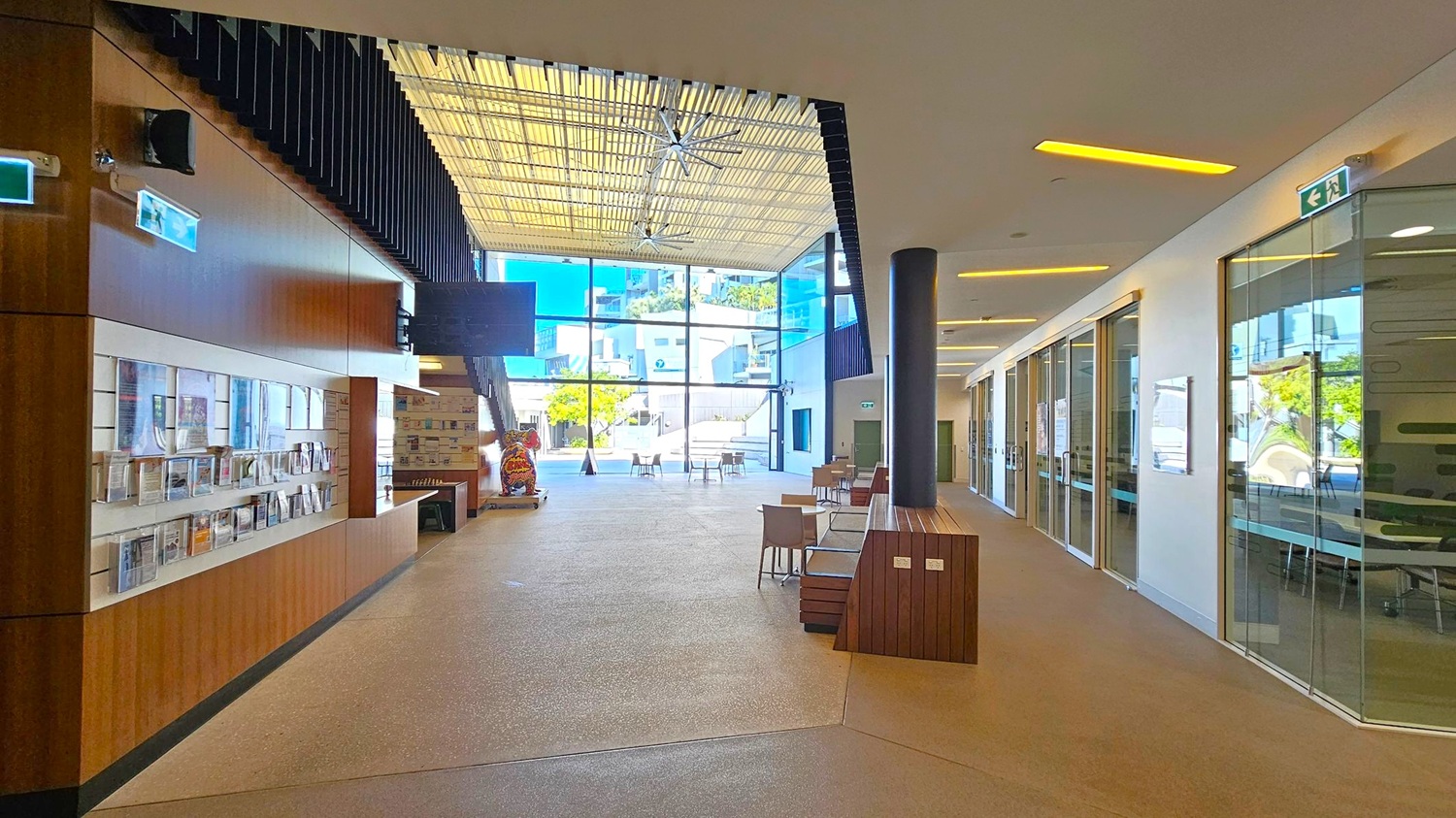 A spacious interior with a high ceiling featuring a large fan. On the left, there is a wooden wall with shelves displaying brochures and magazines. The right side has glass-walled rooms with visible tables and chairs inside. In the centre, there are tables and chairs arranged in an open area. The background shows large glass windows with a view of a courtyard and buildings outside. The floor is a light-coloured polished surface. A spacious interior with a high ceiling featuring a large fan. On the left, there is a wooden wall with shelves displaying brochures and magazines. The right side has glass-walled rooms with visible tables and chairs inside. In the centre, there are tables and chairs arranged in an open area. The background shows large glass windows with a view of a courtyard and buildings outside. The floor is a light-coloured polished surface.