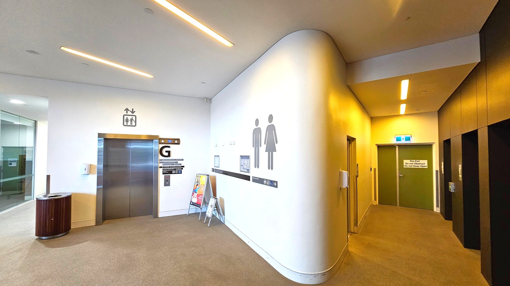 A corridor with a beige carpeted floor and a white ceiling featuring recessed lighting. On the left, there's a lift with a "G" sign above it, a bin nearby, and a glass wall. In the centre, a curved wall displays male and female restroom symbols. On the right, a hallway leads to a green door marked as a fire exit. The walls are light-coloured, and there are additional lifts with dark panels on the far right. A corridor with a beige carpeted floor and a white ceiling featuring recessed lighting. On the left, there's a lift with a "G" sign above it, a bin nearby, and a glass wall. In the centre, a curved wall displays male and female restroom symbols. On the right, a hallway leads to a green door marked as a fire exit. The walls are light-coloured, and there are additional lifts with dark panels on the far right.