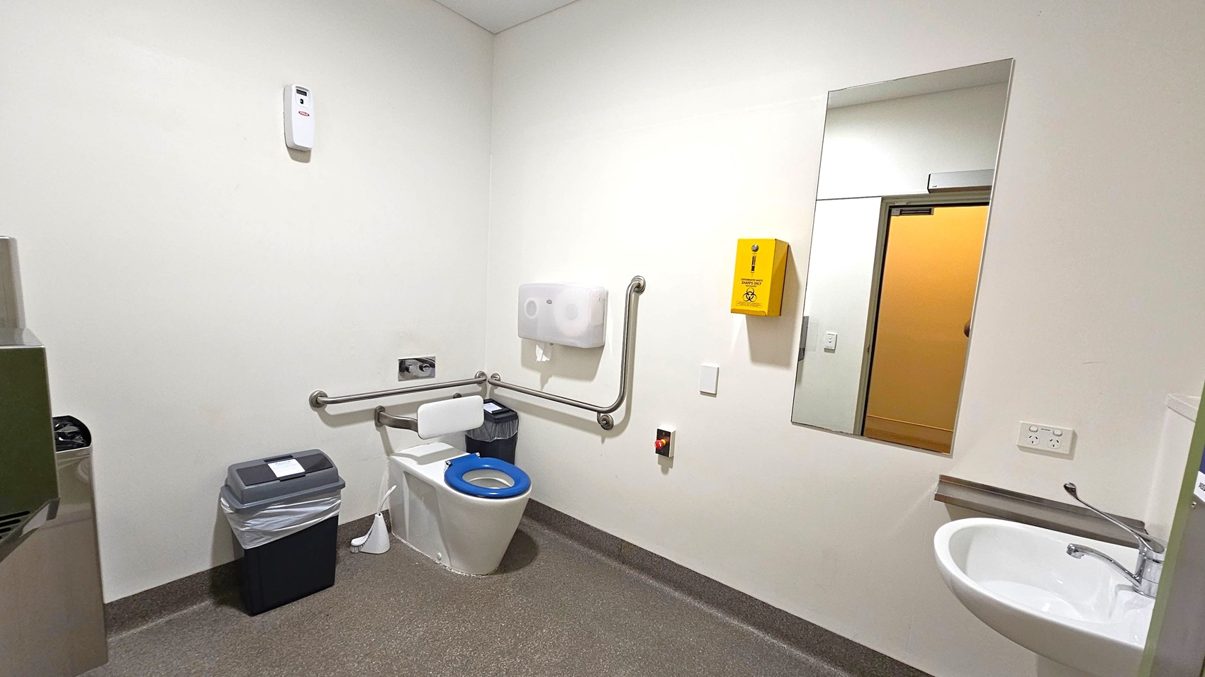 A clean, accessible bathroom with a white toilet featuring a blue seat and support rails on the left. A bin with a liner is next to the toilet. Above, a paper dispenser and a yellow sharps disposal box are mounted on the wall. On the right, a large mirror reflects the opposite wall and a door. Below the mirror, a white sink with a lever tap is installed. The floor is grey with a textured surface, and the walls are plain white. A clean, accessible bathroom with a white toilet featuring a blue seat and support rails on the left. A bin with a liner is next to the toilet. Above, a paper dispenser and a yellow sharps disposal box are mounted on the wall. On the right, a large mirror reflects the opposite wall and a door. Below the mirror, a white sink with a lever tap is installed. The floor is grey with a textured surface, and the walls are plain white.