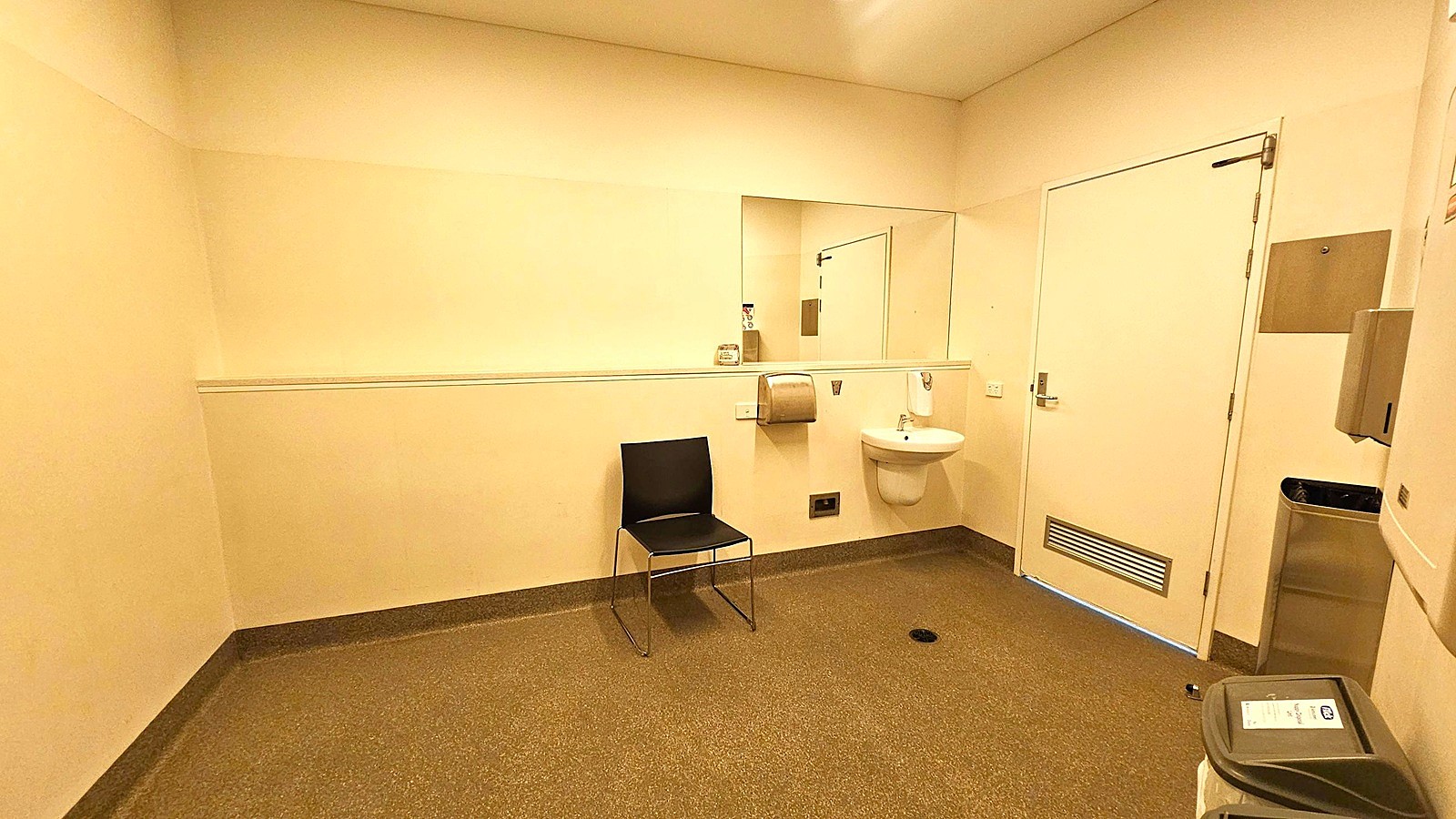 A spacious, beige-walled room with a speckled grey floor. On the right, a white door with a vent at the bottom. Next to it, a small white sink with a soap dispenser above and a paper towel dispenser to the left. A black chair is positioned against the wall beneath a large mirror. In the foreground, a grey bin with a lid. The ceiling is plain with recessed lighting. A spacious, beige-walled room with a speckled grey floor. On the right, a white door with a vent at the bottom. Next to it, a small white sink with a soap dispenser above and a paper towel dispenser to the left. A black chair is positioned against the wall beneath a large mirror. In the foreground, a grey bin with a lid. The ceiling is plain with recessed lighting.