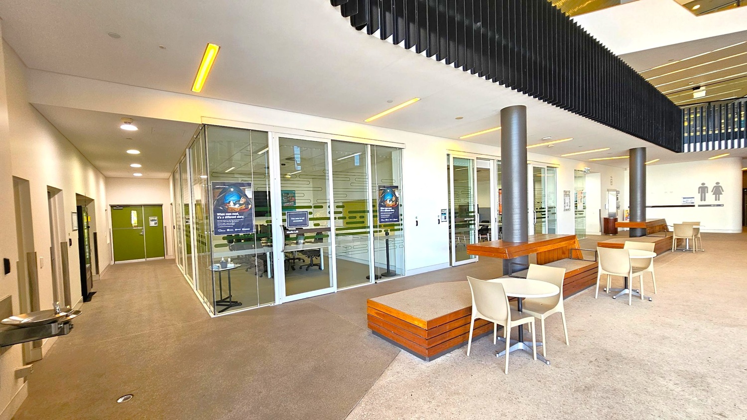 A modern indoor space with a beige carpeted floor. On the left, a corridor with green double doors and a water fountain. The middle features glass walls with posters, revealing a room with tables and chairs. On the right, there are round tables with beige chairs and wooden benches around black pillars. The ceiling has recessed lighting and black slatted panels. Restroom signs are visible in the background. A modern indoor space with a beige carpeted floor. On the left, a corridor with green double doors and a water fountain. The middle features glass walls with posters, revealing a room with tables and chairs. On the right, there are round tables with beige chairs and wooden benches around black pillars. The ceiling has recessed lighting and black slatted panels. Restroom signs are visible in the background.