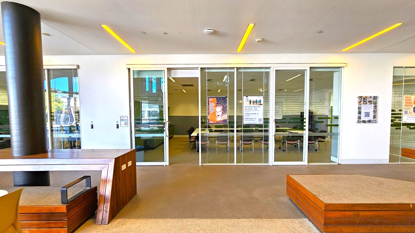 A modern interior space with a glass-walled room in the centre, featuring sliding doors. Inside, there are tables and chairs arranged in a meeting setup. Posters are visible on the glass, with one showing a smiling child. On the left, a dark cylindrical column and a wooden bench with a metal handle. On the right, a wall-mounted display with a QR code. The ceiling has recessed lighting, and the floor is carpeted in a neutral tone. A modern interior space with a glass-walled room in the centre, featuring sliding doors. Inside, there are tables and chairs arranged in a meeting setup. Posters are visible on the glass, with one showing a smiling child. On the left, a dark cylindrical column and a wooden bench with a metal handle. On the right, a wall-mounted display with a QR code. The ceiling has recessed lighting, and the floor is carpeted in a neutral tone.